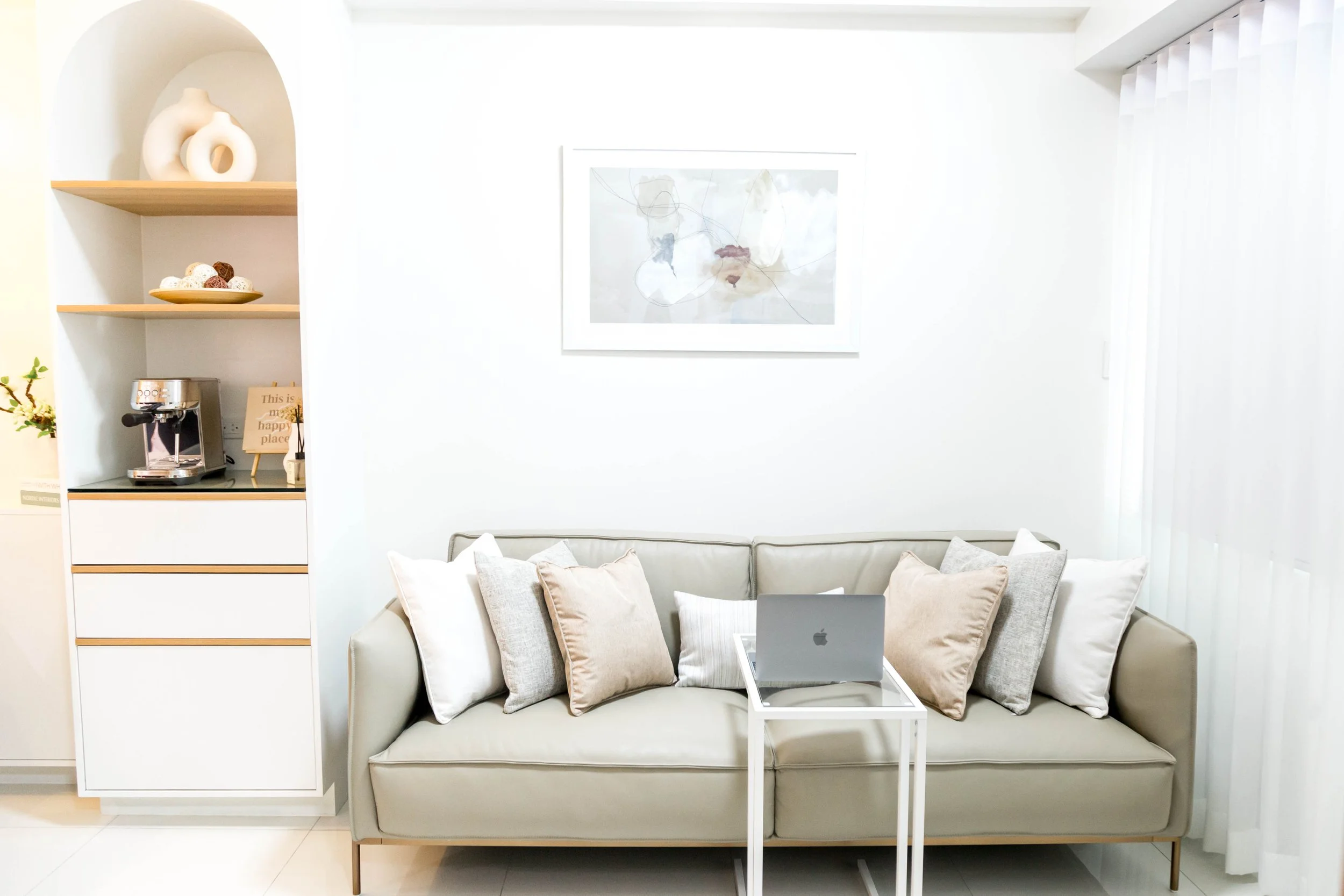 A modern, white living room featuring a beige sofa with multiple cushions, a small white side table with an open laptop, a framed abstract art piece on the wall, built-in shelves with decorative items, a coffee machine, and white curtains.