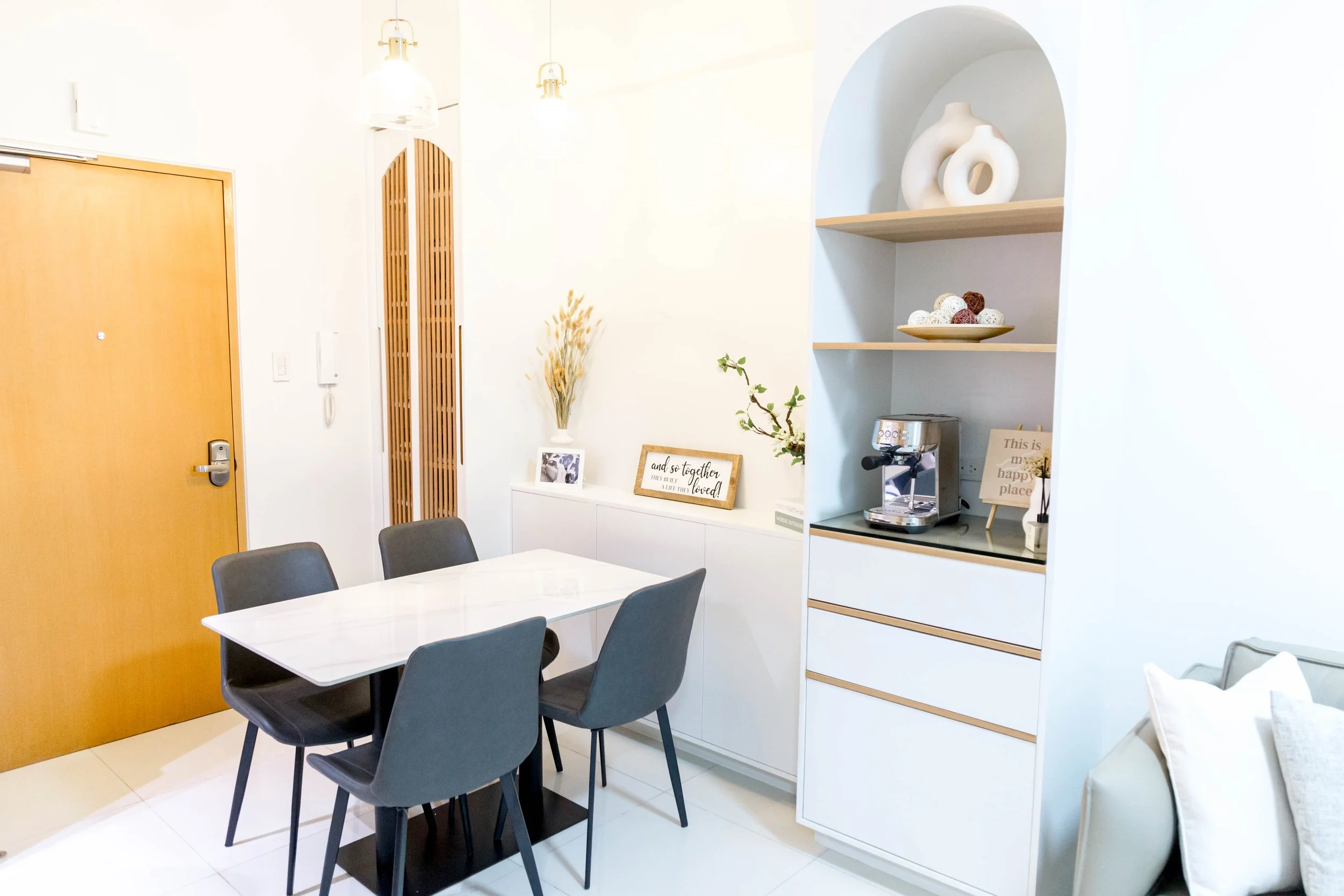 Modern dining area with a white table, six gray chairs, a wooden door, and decorative shelves with vases and signs