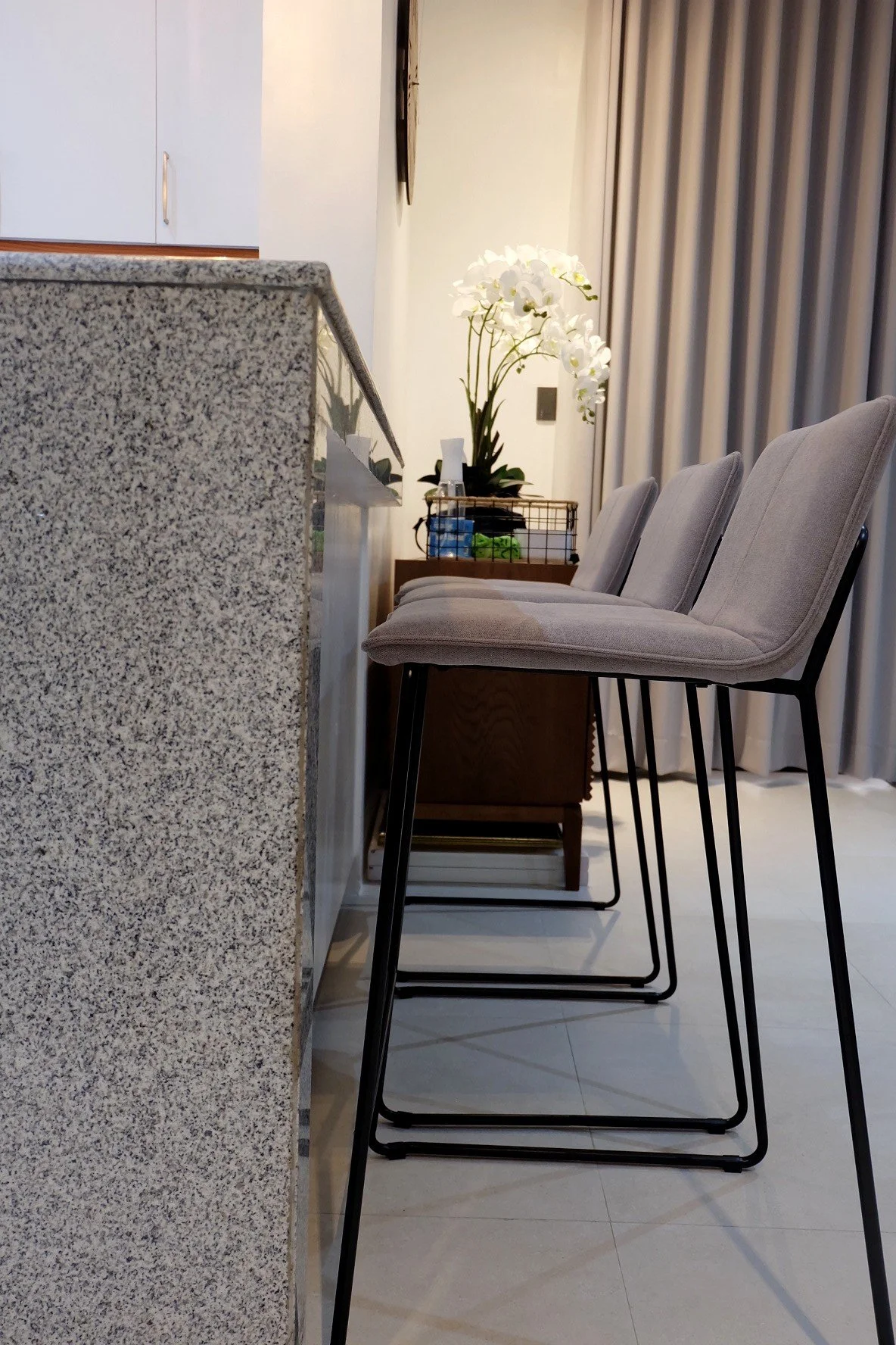 Side view of a kitchen island with three beige upholstered barstools, a granite countertop, and a background featuring a white orchid plant on a wooden side table and a curtain.
