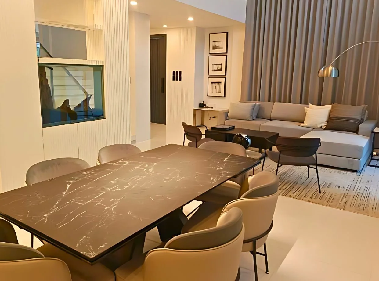 Modern living room and dining area with beige couch, black coffee table, marble dining table with nine chairs, and floor-to-ceiling curtains.
