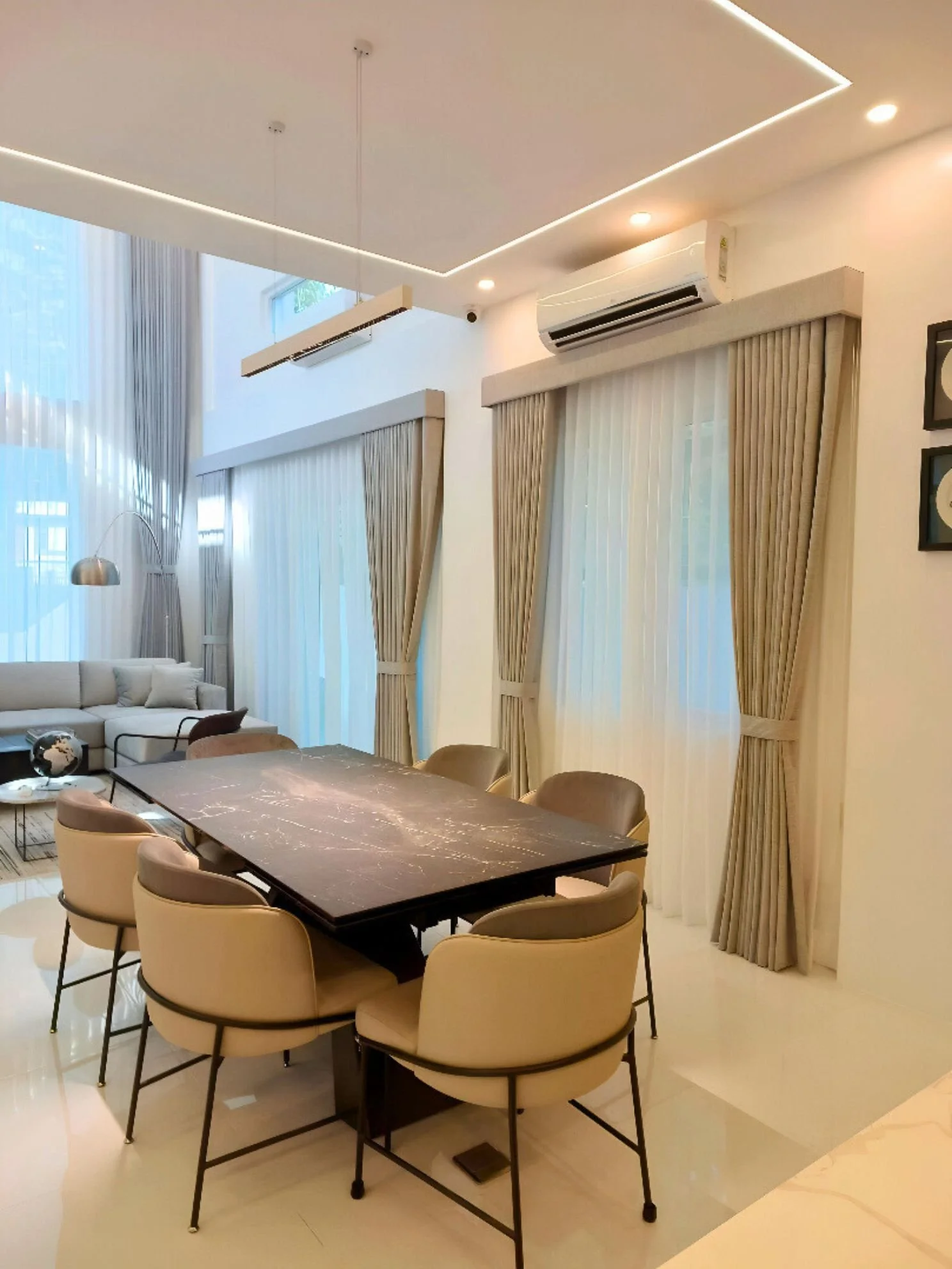 Modern dining room with a large black marble table surrounded by beige chairs, beige curtains covering tall windows, a living area with a sofa and coffee table, and ceiling with recessed lighting.
