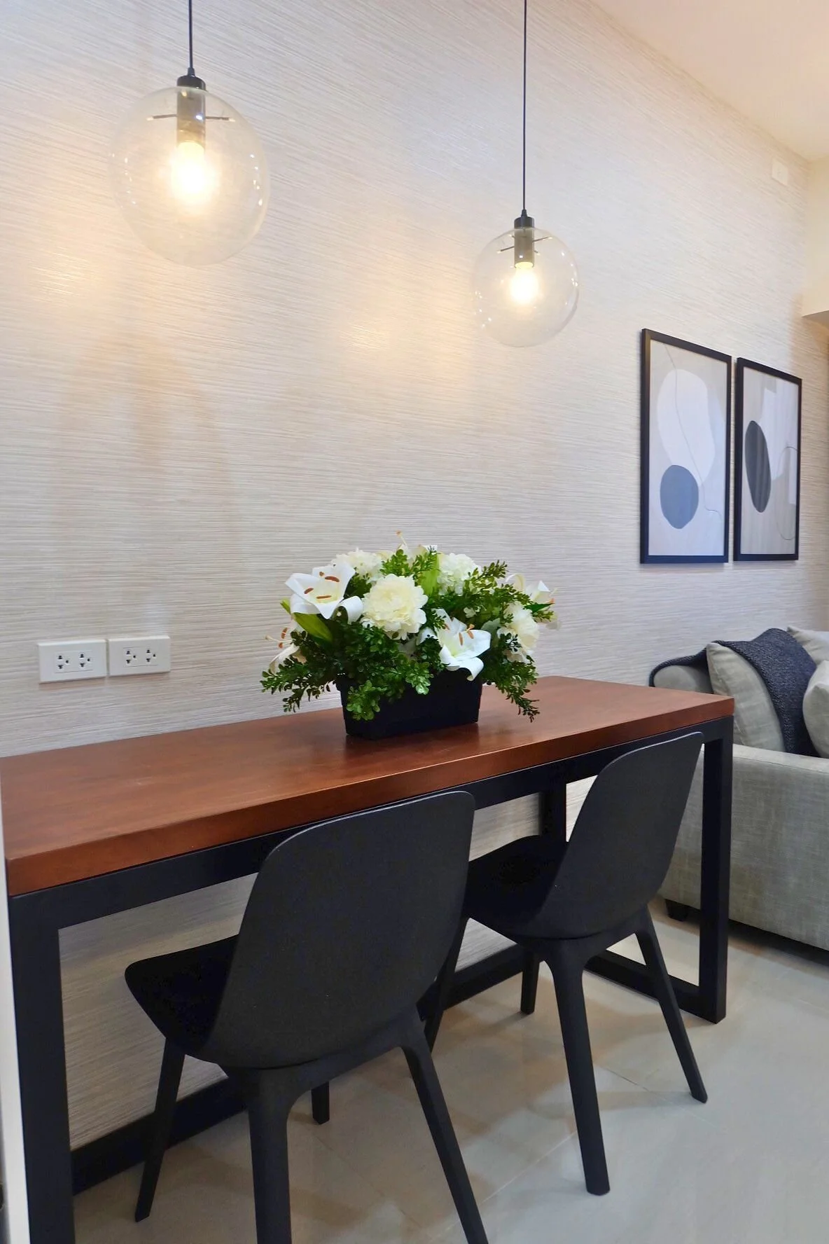 A modern living room corner featuring a wooden table with a floral arrangement, two black chairs, a beige sofa with pillows, and three hanging pendant lights with glass globes. Two framed abstract art pieces are hung on the wall.