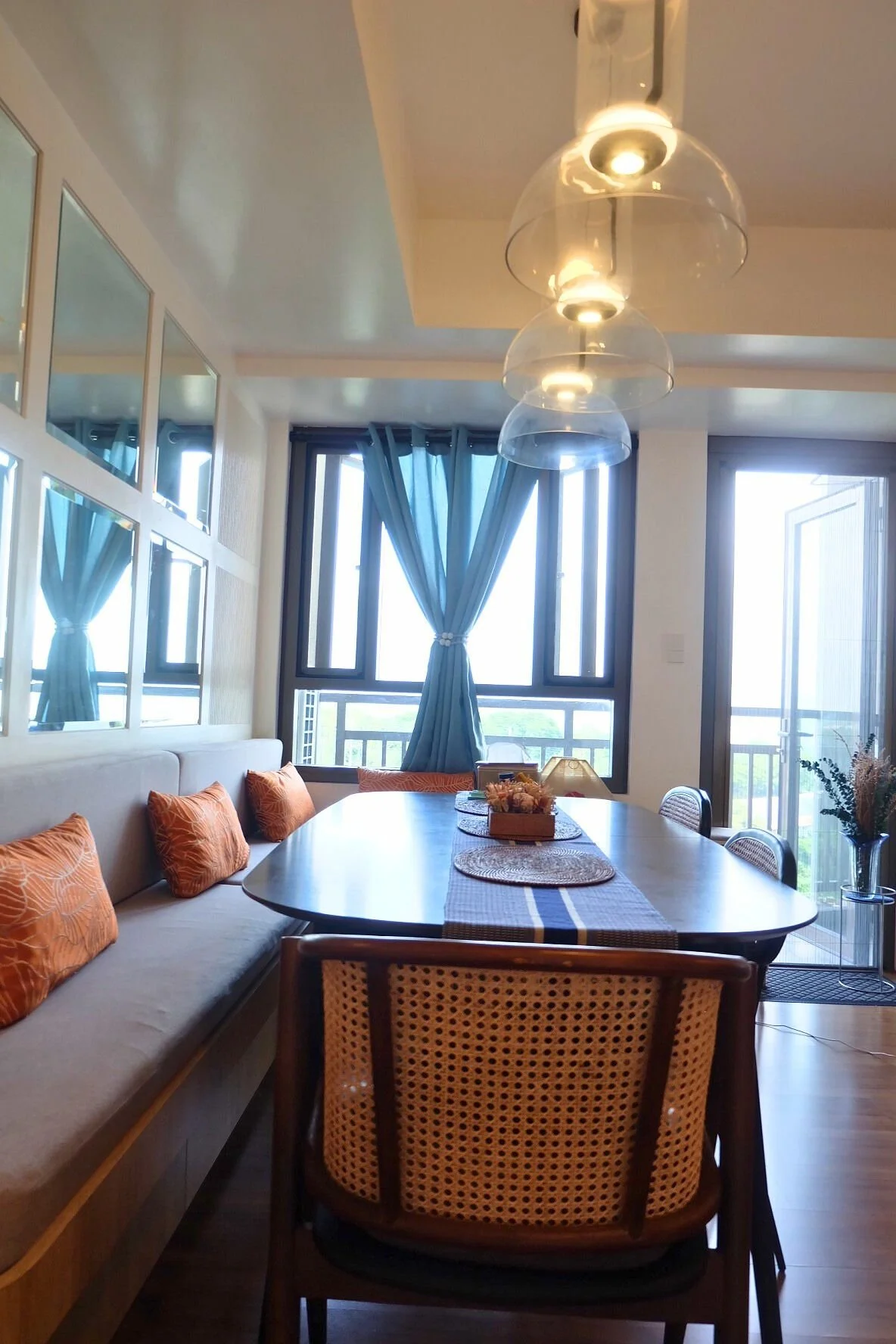 A modern dining room with a wooden table, orange pillows on a bench, a rattan chair, and large windows with blue curtains. A ceiling light fixture with three glass shades hangs above the table, and there is a view of a balcony outside.