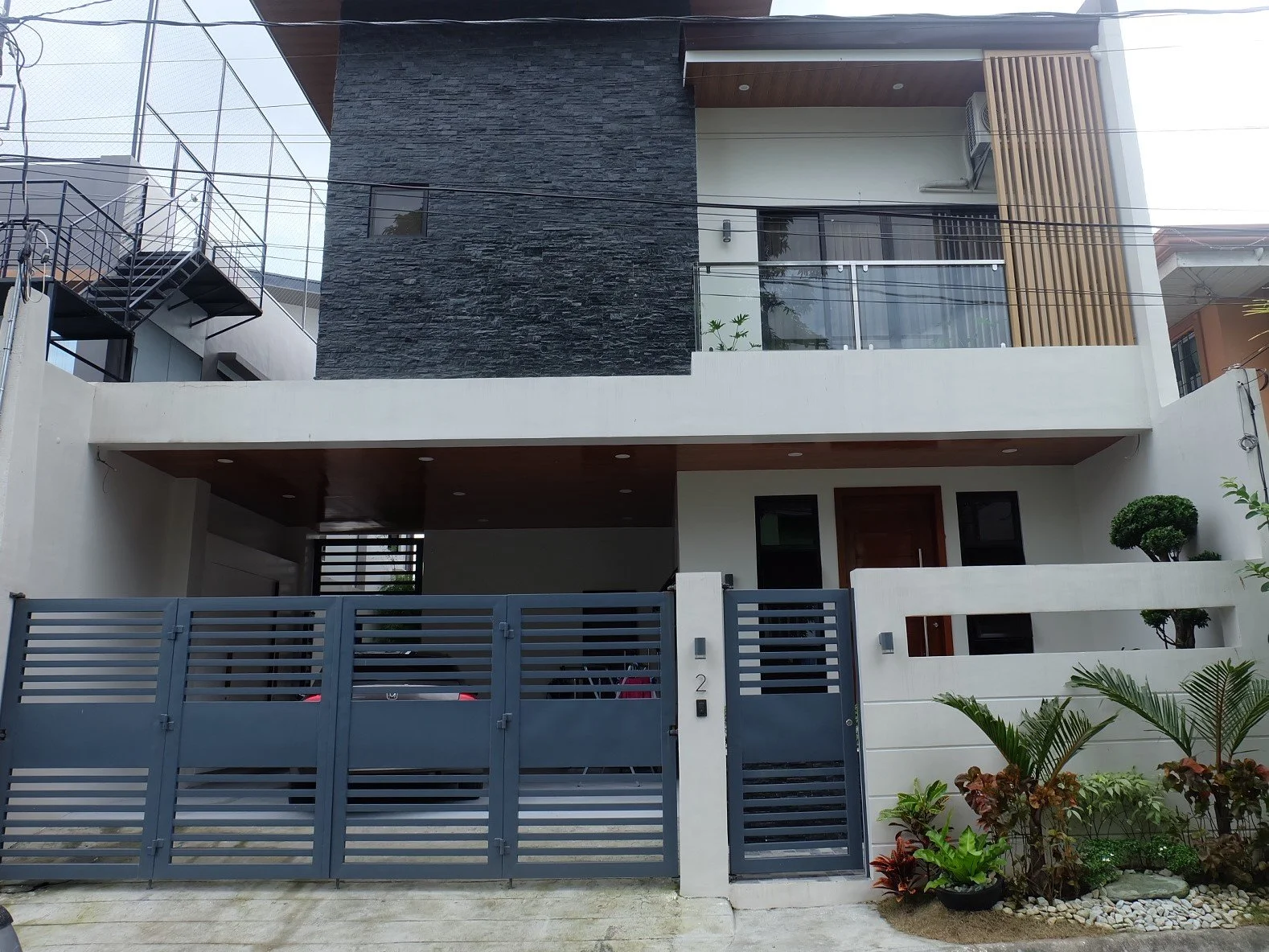 Modern multi-story house with a gray metal gate, white walls, a balcony with glass railing, and decorative plants outside.
