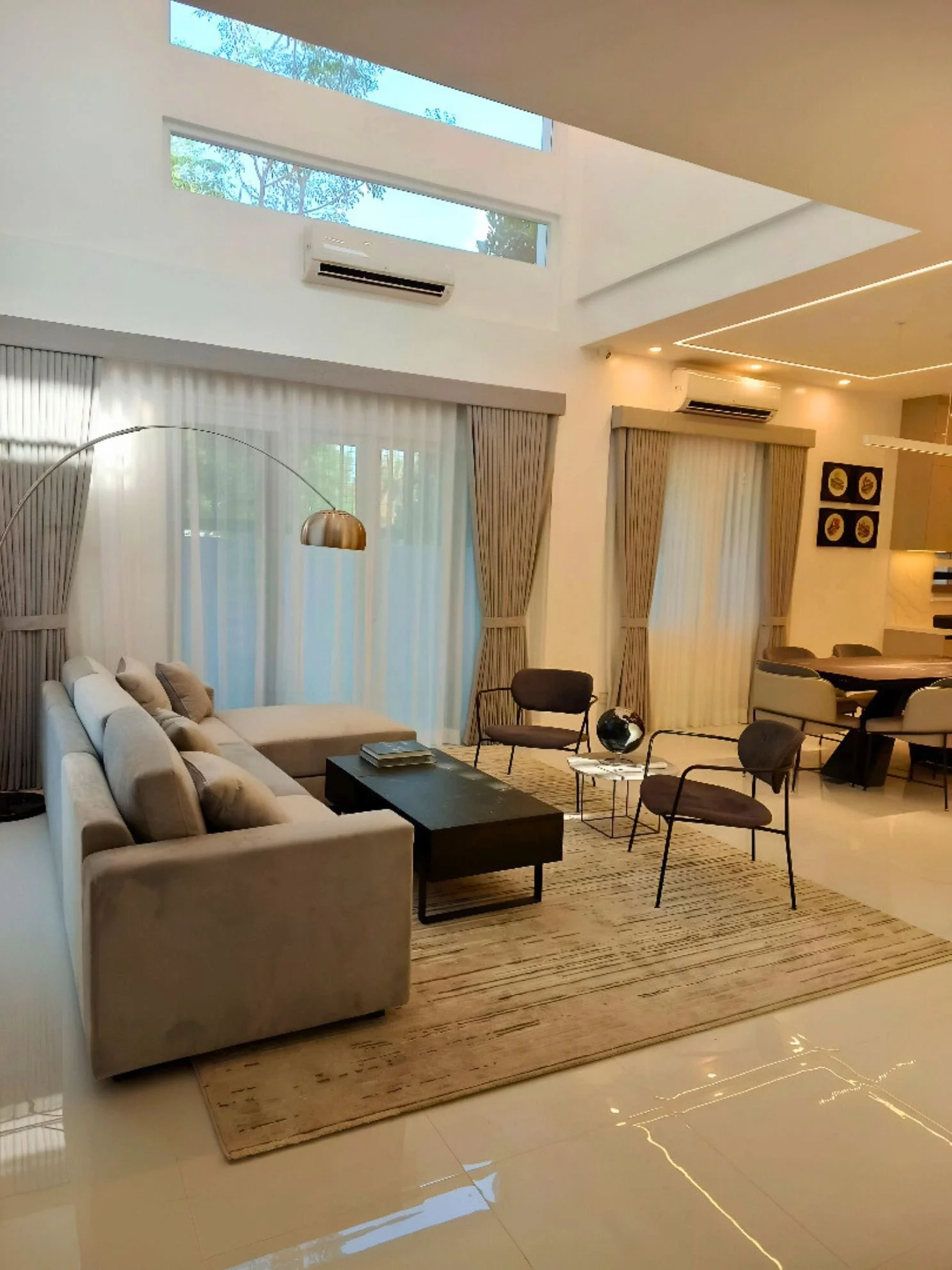 Modern living room with beige sectional sofa, black coffee table, and three brown chairs. Large windows with sheer white curtains and framed artwork on the wall. Skylights and recessed lighting illuminate the space.