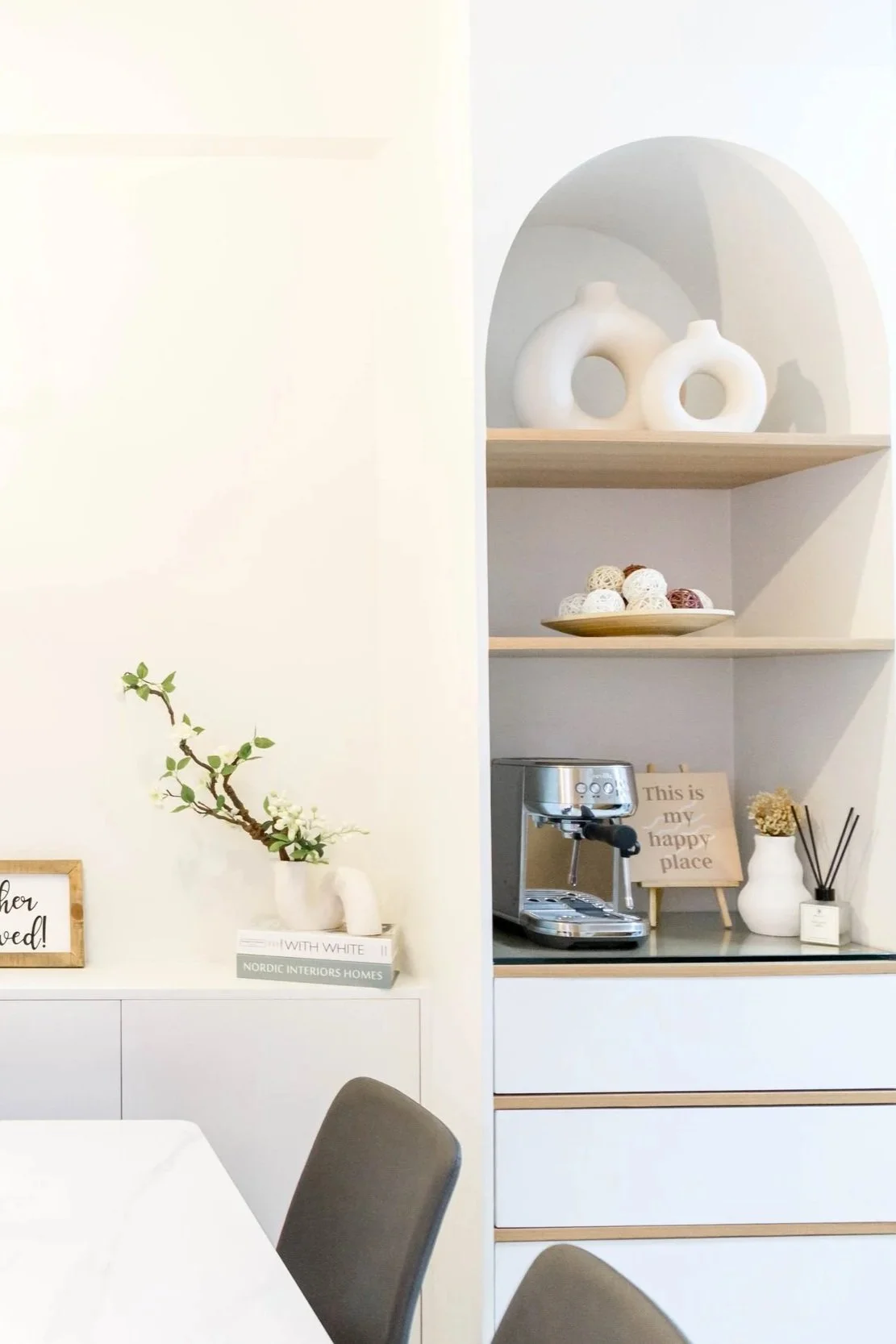 Modern dining area with white walls, a white table, black chairs, decorative vases, a small cabinet with a sign reading 'This is my happy place', shelves with white and wooden vases, a book, a coffee machine, and decorative items.