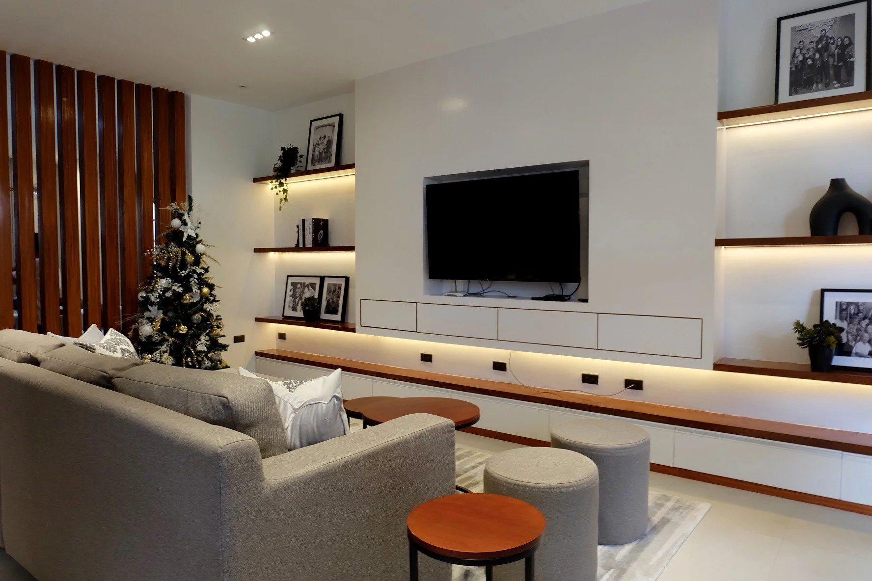 Living room with a beige sofa, a small round wooden coffee table, two beige stools, a decorated Christmas tree, wall-mounted black and white photographs, and a flat-screen TV on a white wall with built-in shelves and warm lighting.