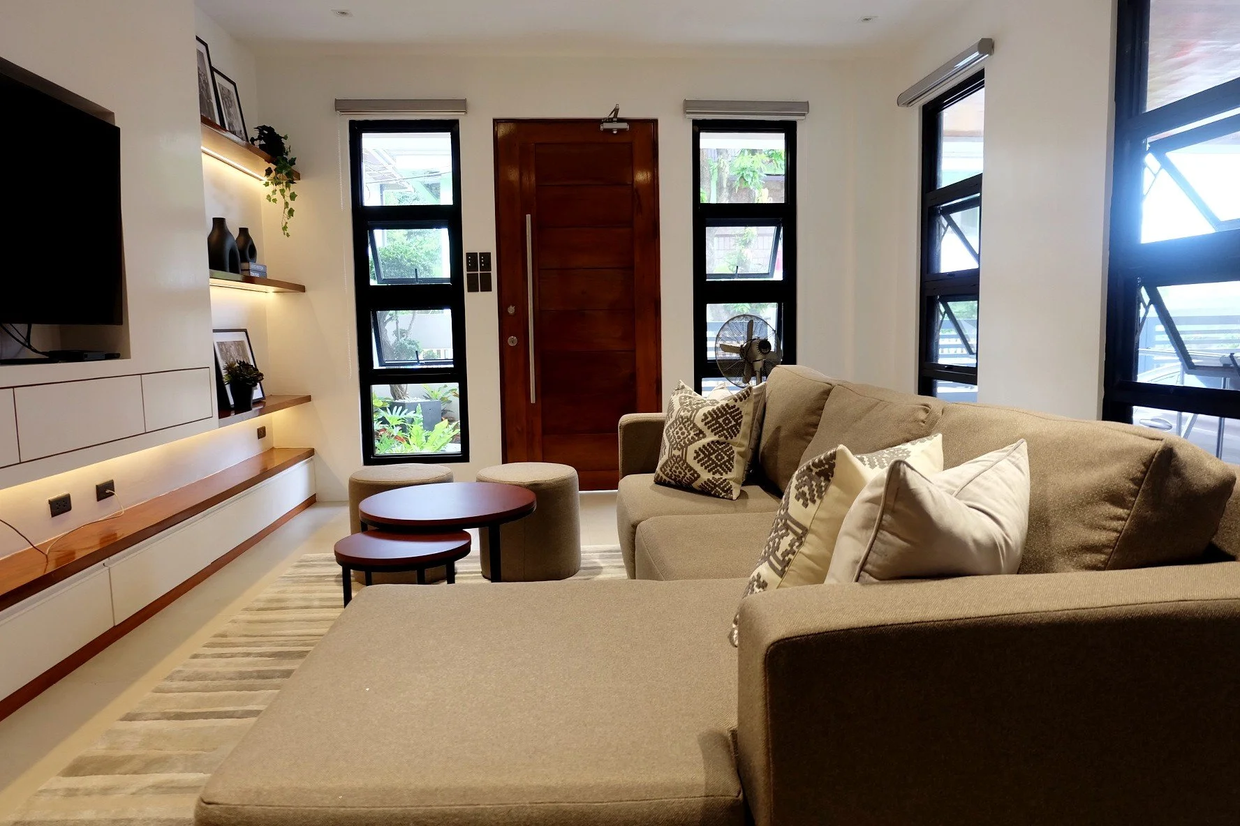 Living room with beige sectional sofa with patterned pillows, small nested round tables, a wall-mounted TV, shelves with decor and photos, front door, and multiple windows with views of outdoor greenery.
