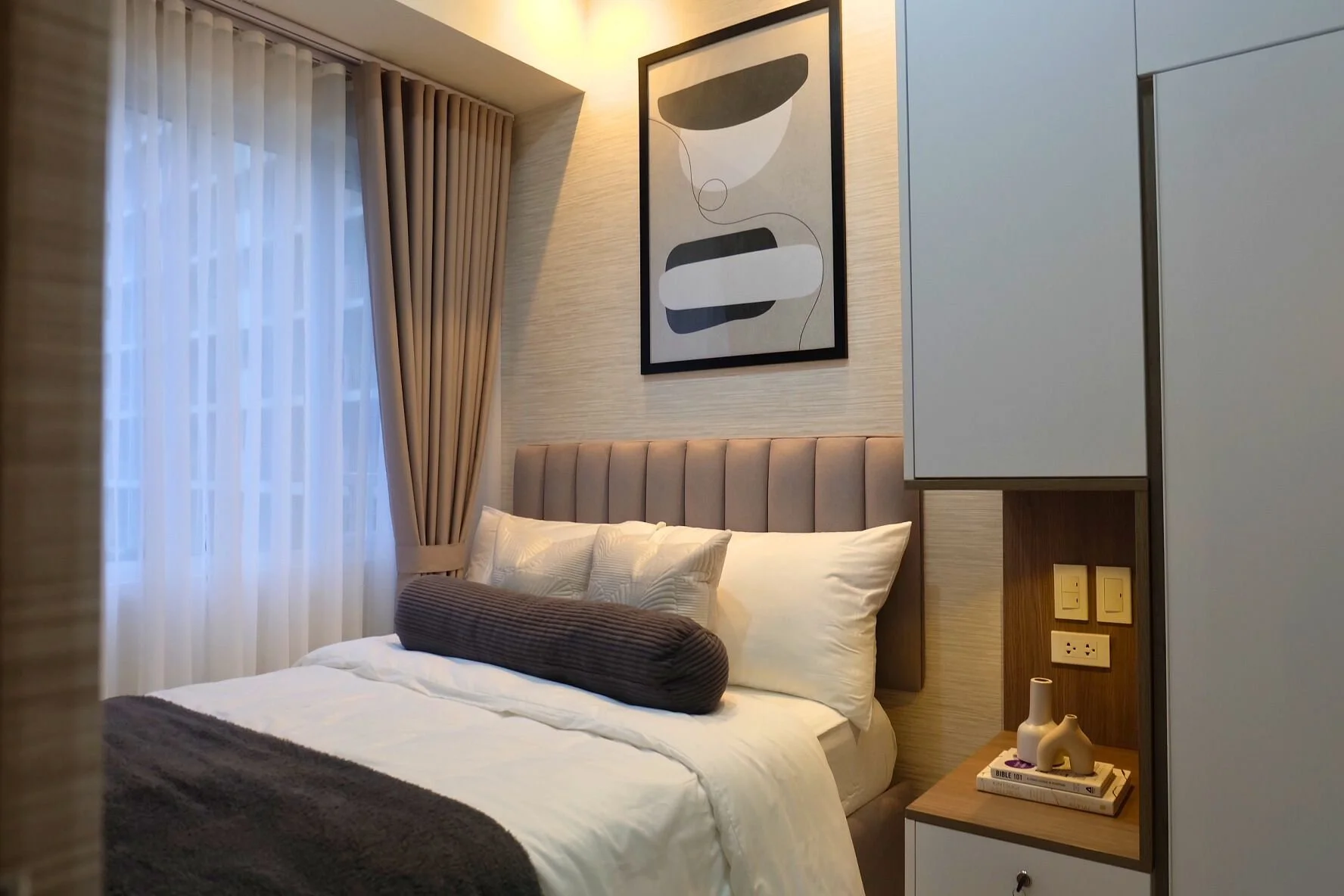 A modern bedroom with a large bed, beige headboard, white bedding, brown decorative bolster pillow, and blanket. There are beige curtains and a window, with an abstract black-and-white wall art piece above the bed. Beside the bed is a small wooden be