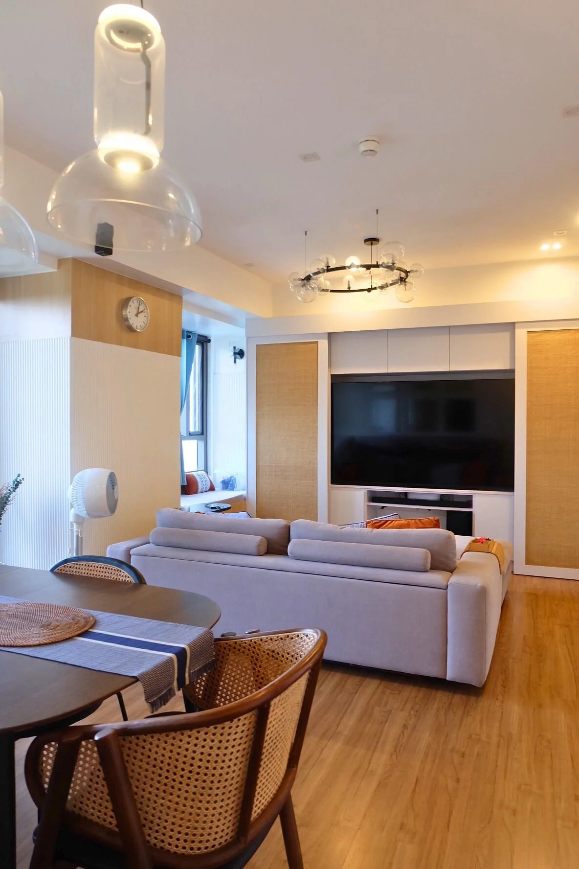 Living room with a beige sofa, TV on the wall, and a dining table with chairs. Modern lighting fixtures and a window with curtains are visible.