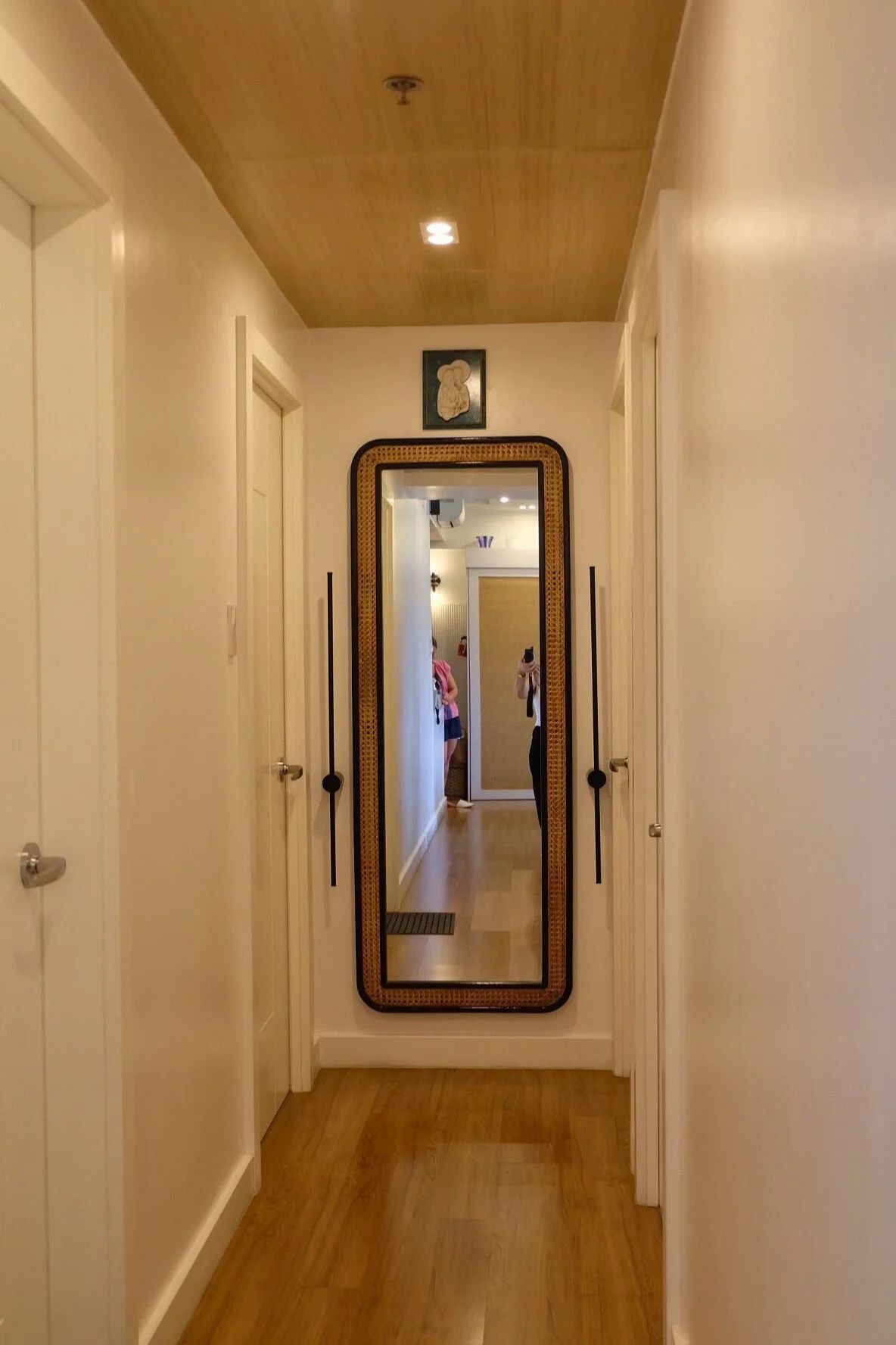 Hallway with wooden ceiling, white walls, and a full-length mirror with a woven frame. Two people are visible in the mirror, one taking a photo and the other standing nearby.