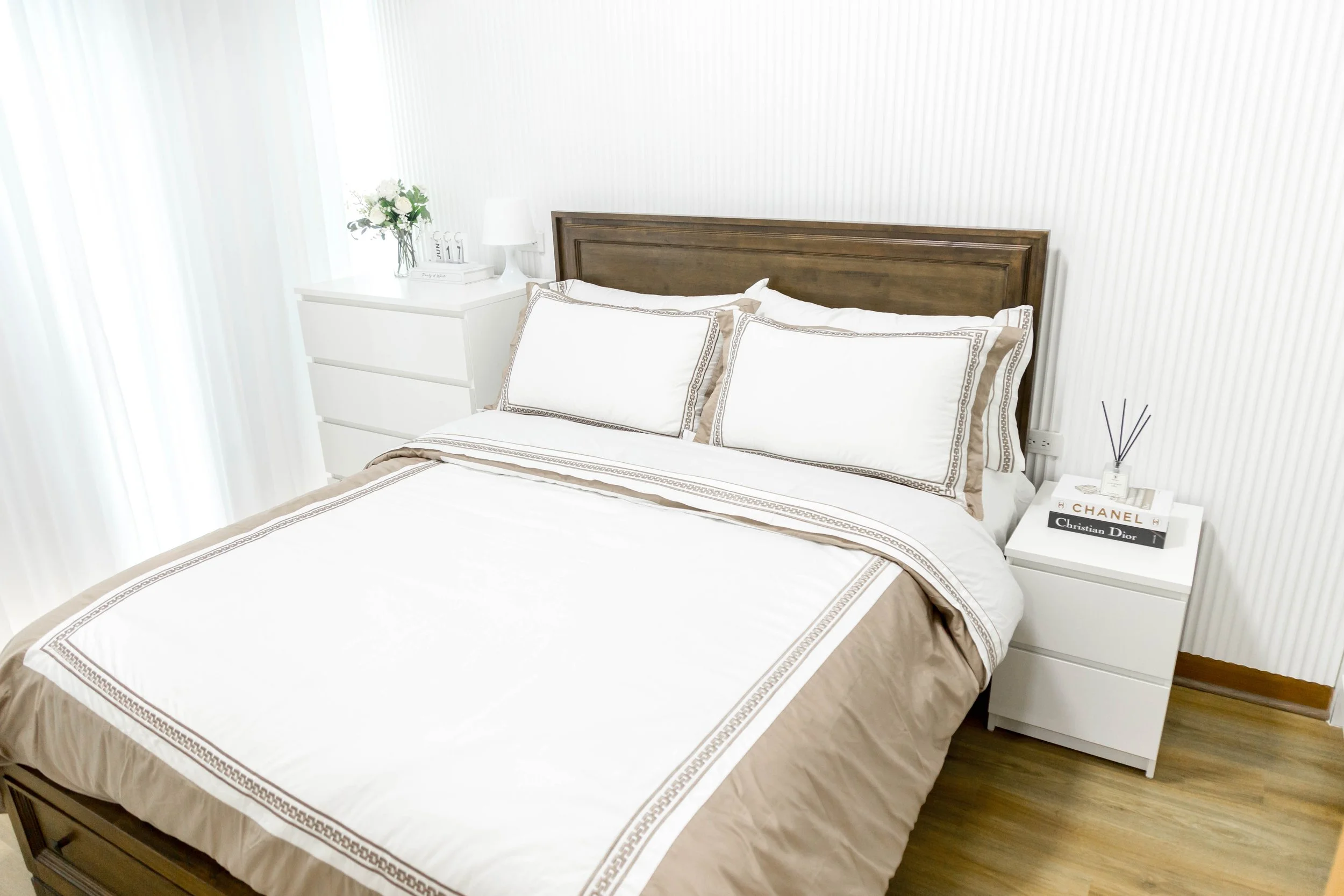 A neatly made bed with a dark wood headboard, white sheets with beige borders, and matching pillowcases in a bright bedroom with a white dresser, a white nightstand, a small lamp, books, and a decorative plant.
