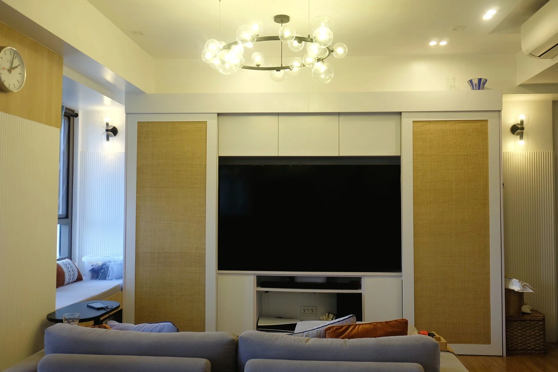 Living room with a large flat-screen TV built into a white entertainment unit, decorative paneling, and a modern chandelier with spherical bulbs. There is a window seat with pillows and a side table with a glass of water. The room has warm lighting a