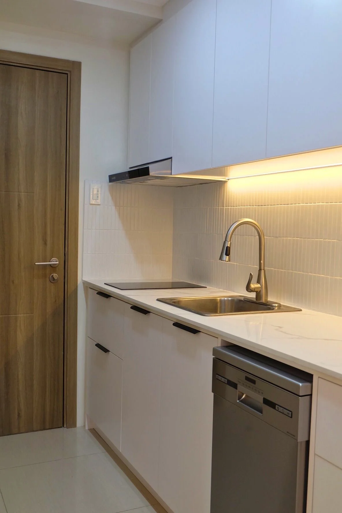 Modern kitchen with white cabinetry, a stainless steel sink with a curved faucet, a built-in electric cooktop, a dishwasher under the counter, beige tiled backsplash, and a wooden door.