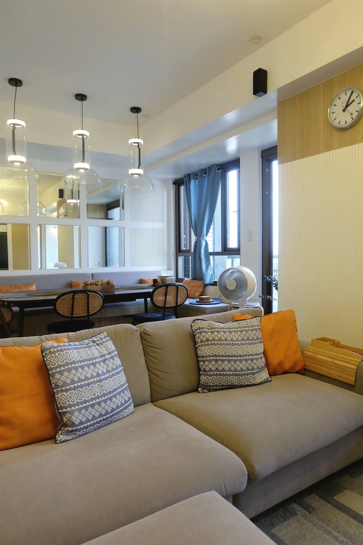 Living room with beige sofa, patterned throw pillows, a wooden wall clock, large windows with blue curtains, a fan on a white stand, a dining table with chairs, and pendant lights hanging from the ceiling.
