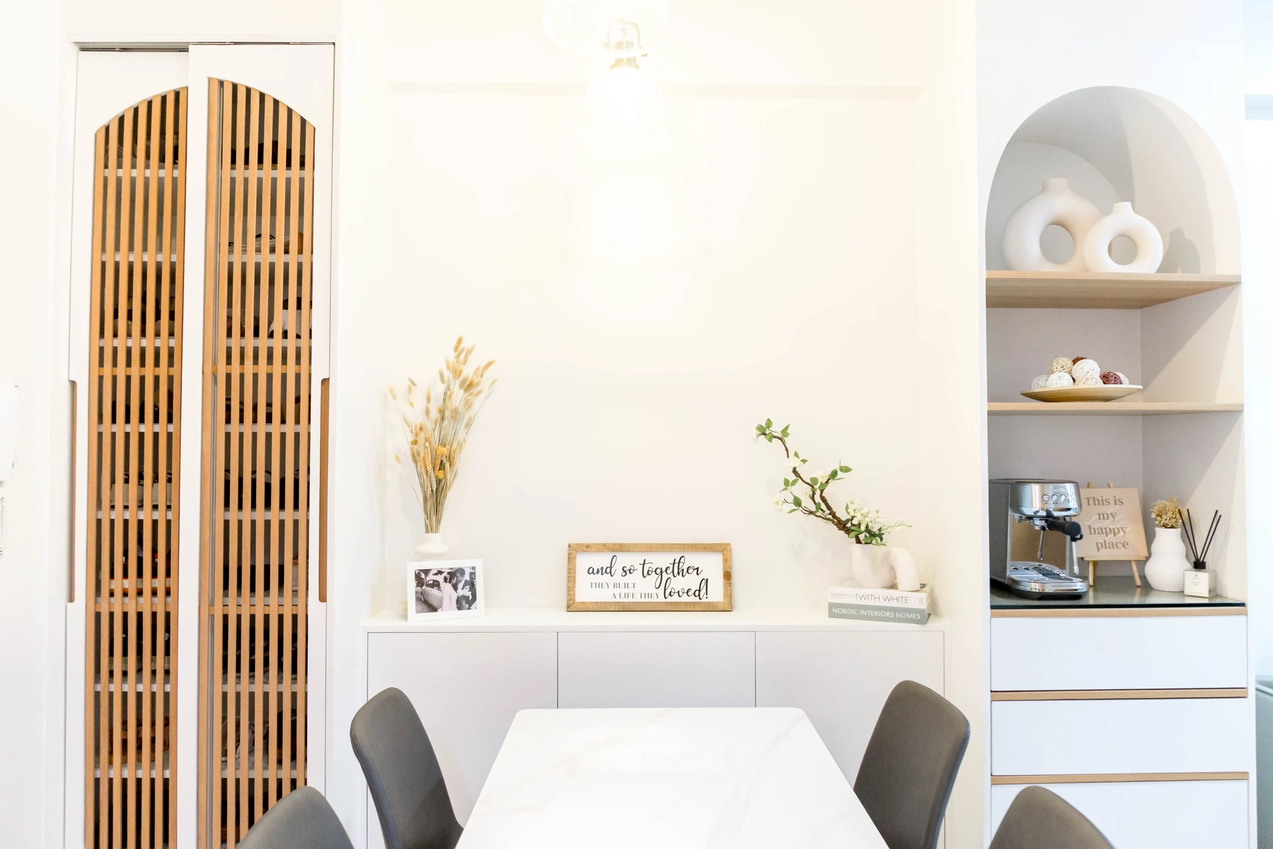 A modern dining area with a white marble table and gray chairs, white decorative shelves with vases and art, a vase with dried flowers, and framed quotes and photos on the wall.
