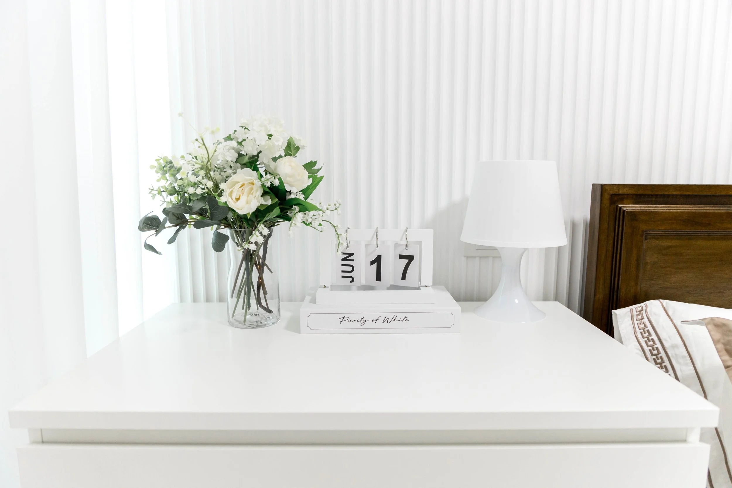 A white bedroom nightstand with a glass vase of white flowers, a white calendar displaying June 17, and a white lamp. A wooden headboard is partially visible on the right.