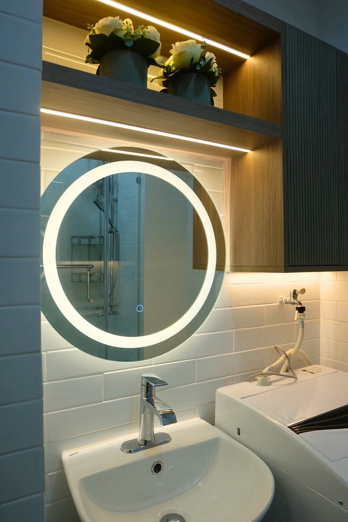 Bathroom with white subway-tile walls, a round backlit mirror, a small white sink with a chrome faucet, a wooden shelf with two potted white flowers above the mirror, shower visible in the reflection, and a white washing machine.