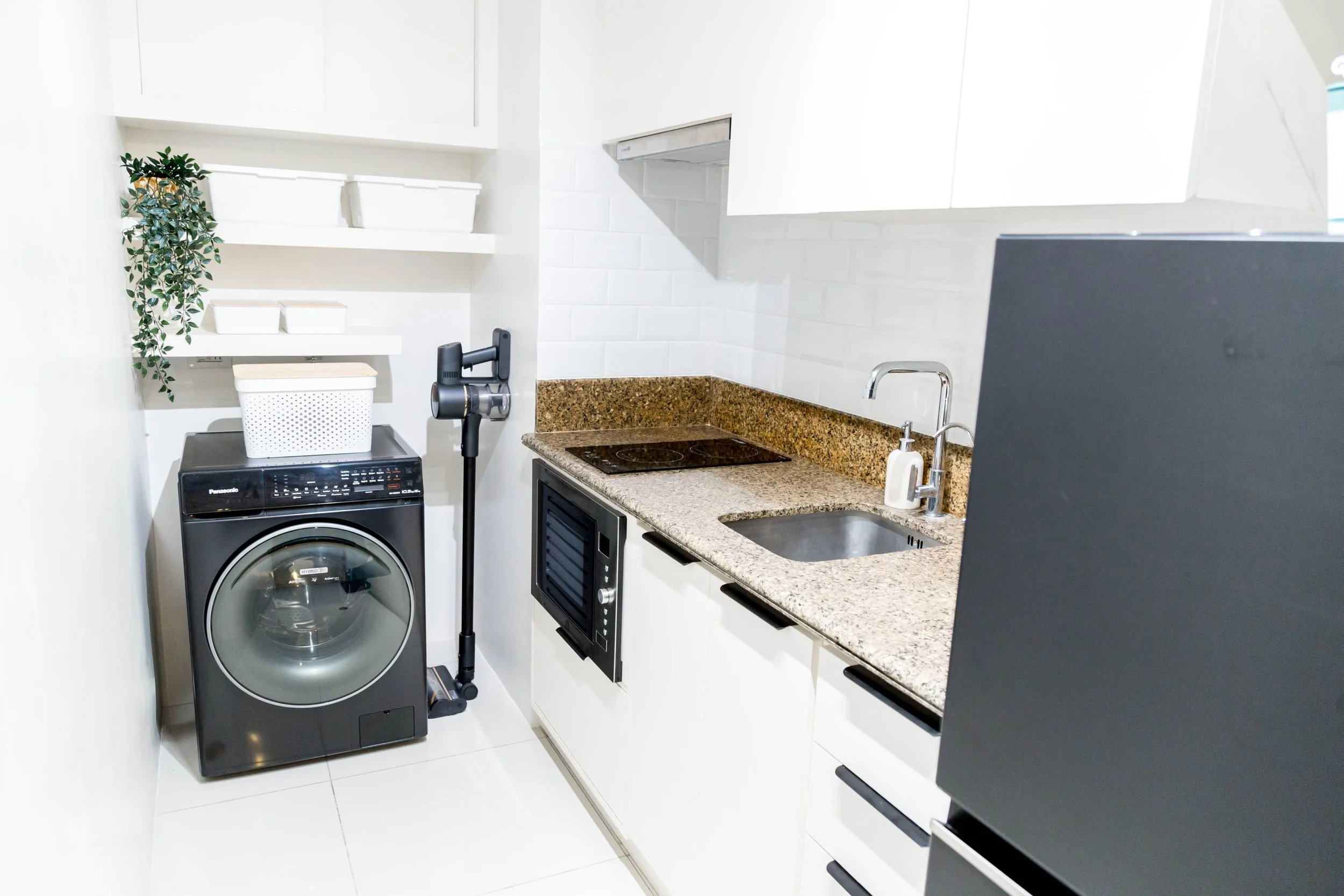 Compact kitchen with white cabinets, granite countertop, stainless steel sink, stovetop, built-in oven, and a washing machine. Open shelves with white containers and a small plant.