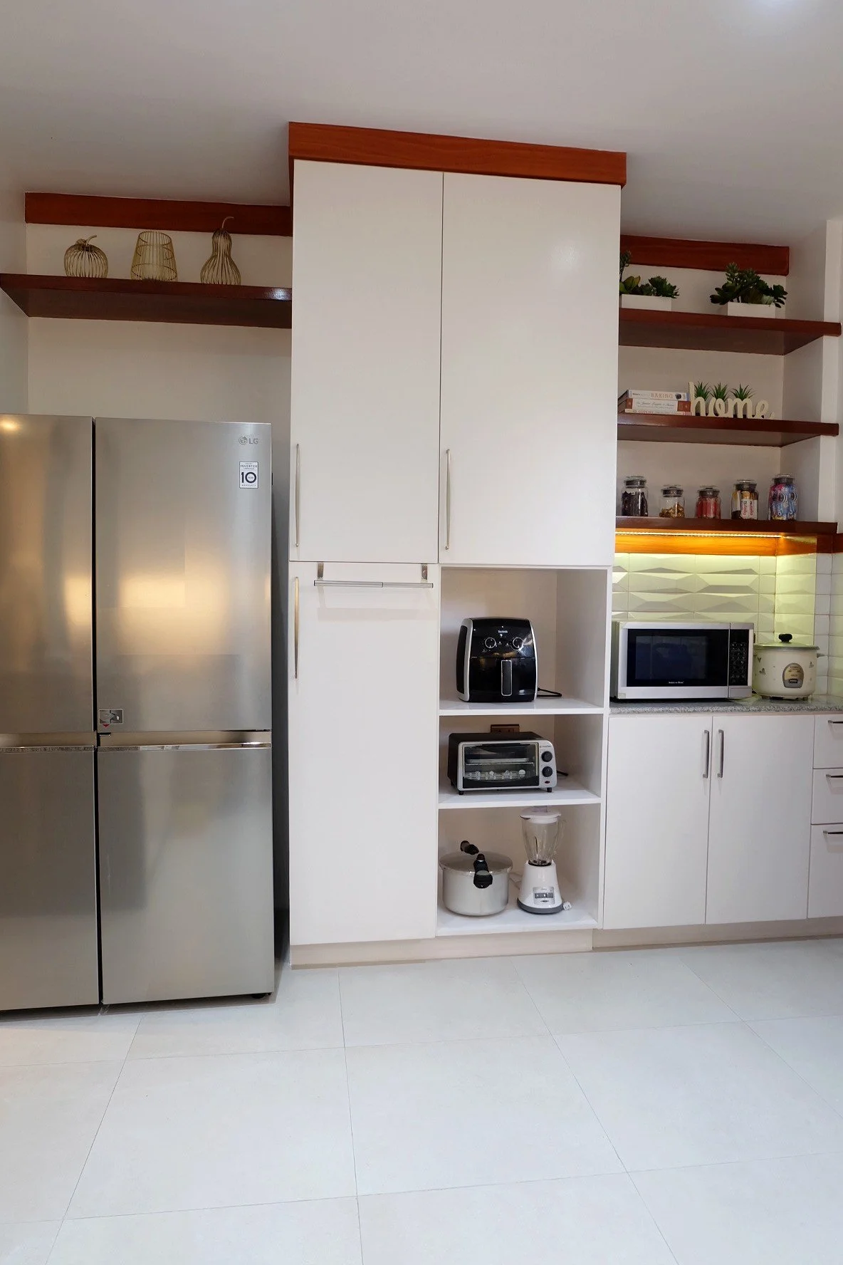 Kitchen with white cabinets, stainless steel refrigerator, and open shelves with decorative items and jars. Counter with microwave, toaster oven, rice cooker, and blender.