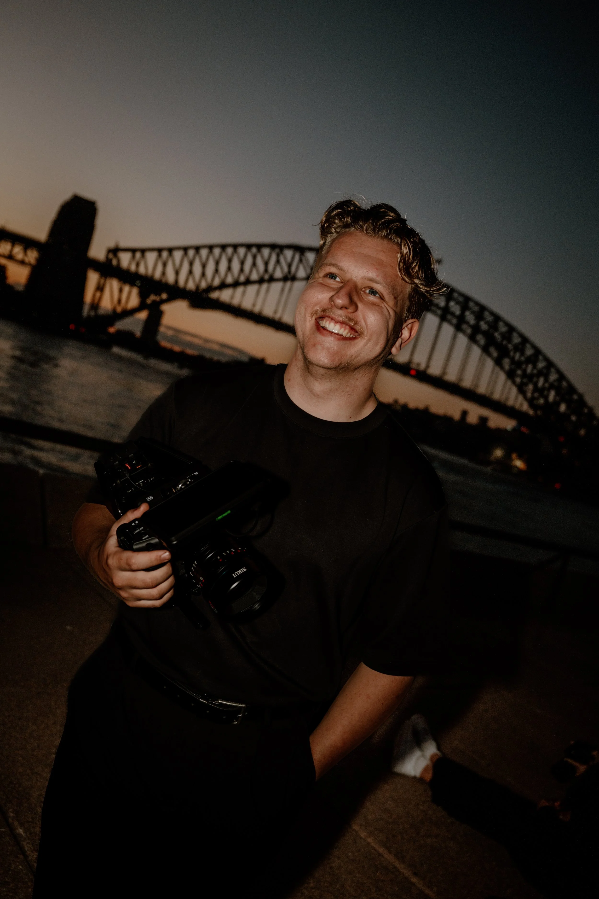 A smiling man holding a black camera with a large lens, standing outside near a bridge during sunset, with a partly cloudy sky.