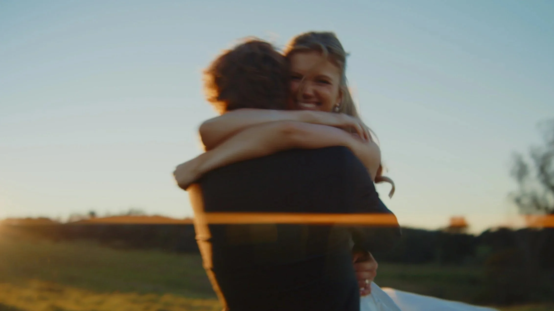 Two people hugging outdoors during sunset, with a landscape and trees in the background.