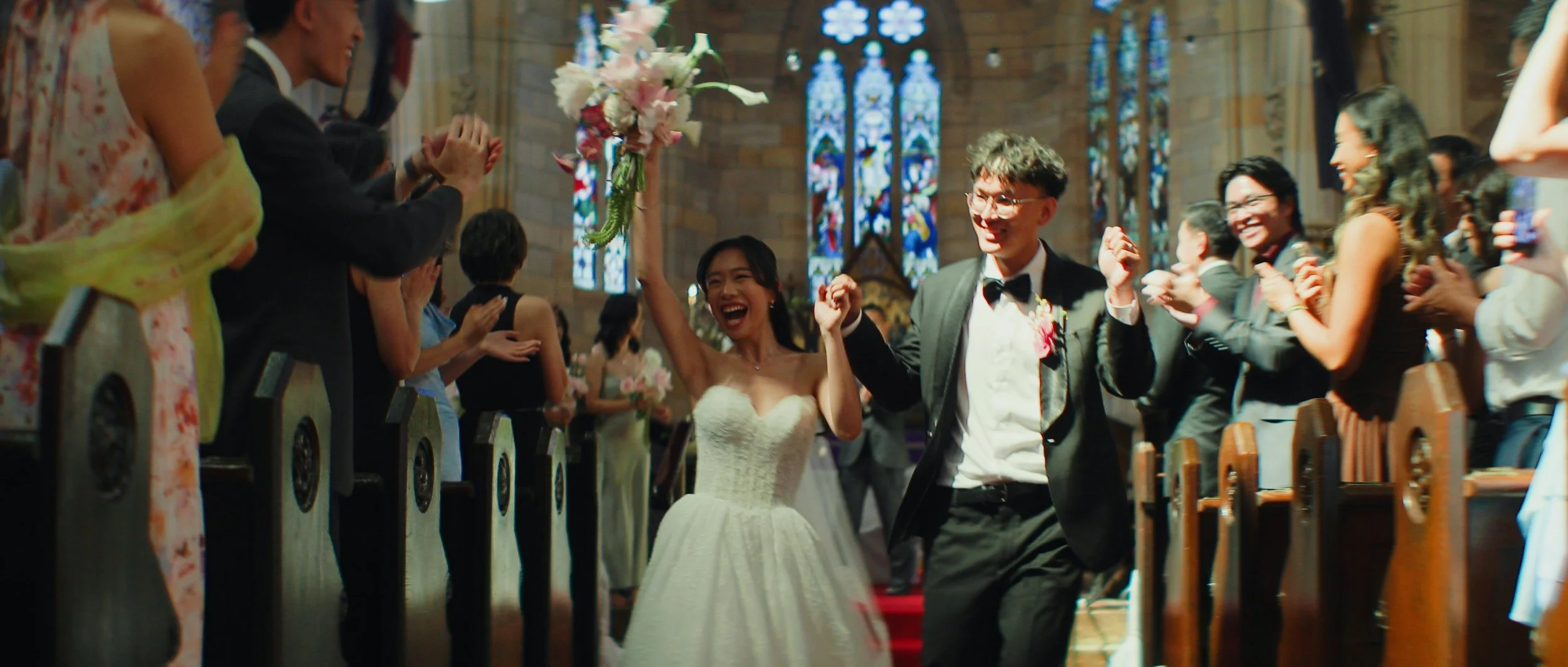 A joyous wedding celebration inside a church, with the bride and groom walking down the aisle holding hands, surrounded by clapping guests, with stained glass windows in the background.