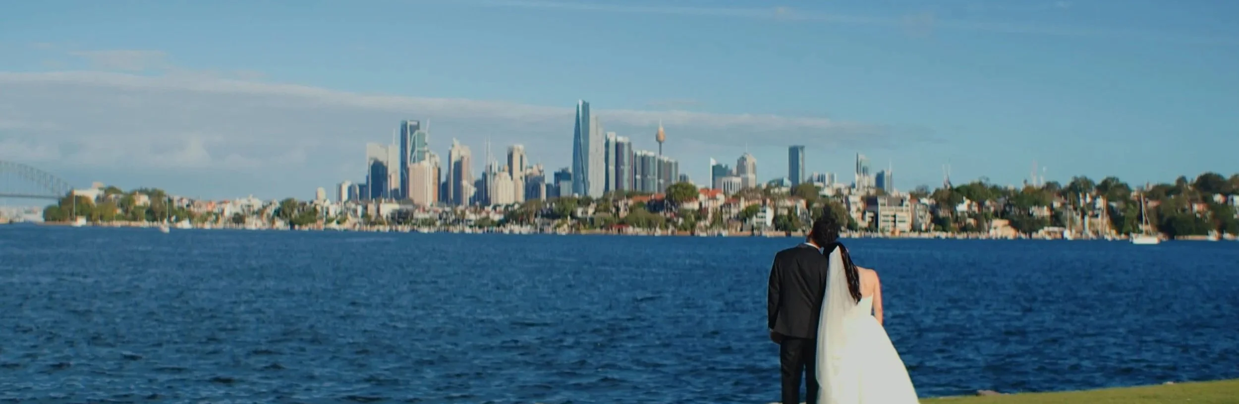 A couple in wedding attire, with a man in a suit and a woman in a white wedding dress, stands by the water with an urban skyline of a city, possibly Sydney, in the background, under a partly cloudy sky.