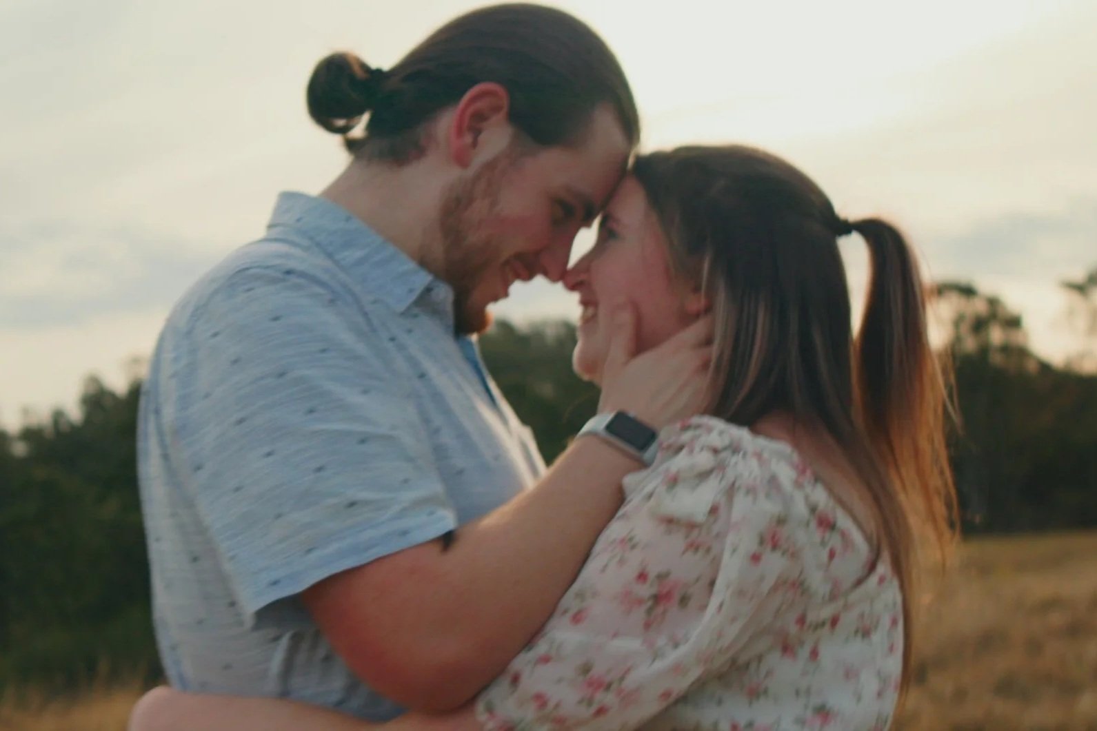 A man and woman are outdoors, touching foreheads and smiling at each other during sunset, with trees in the background.