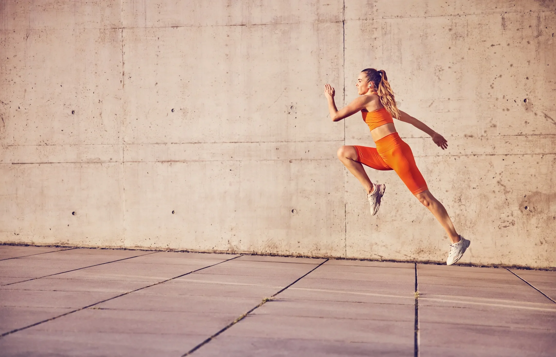 A woman in orange athletic wear is captured mid-air jumping against a plain concrete wall.