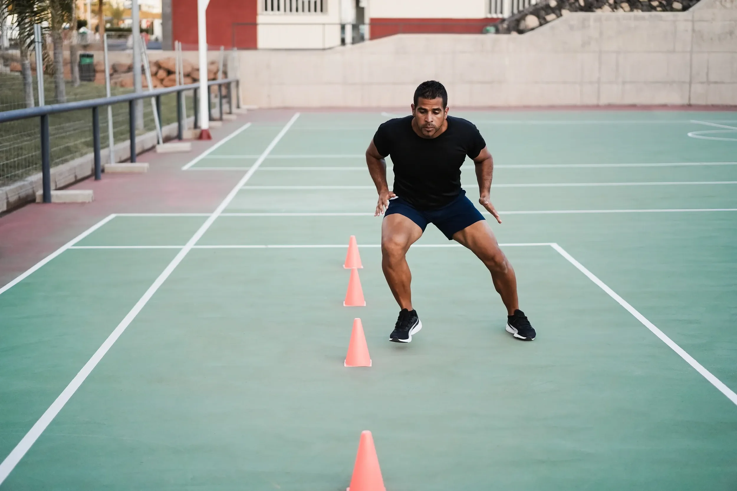 A man wearing a black t-shirt, navy shorts, and black sneakers performs agility drills through orange cones on an outdoor tennis court.