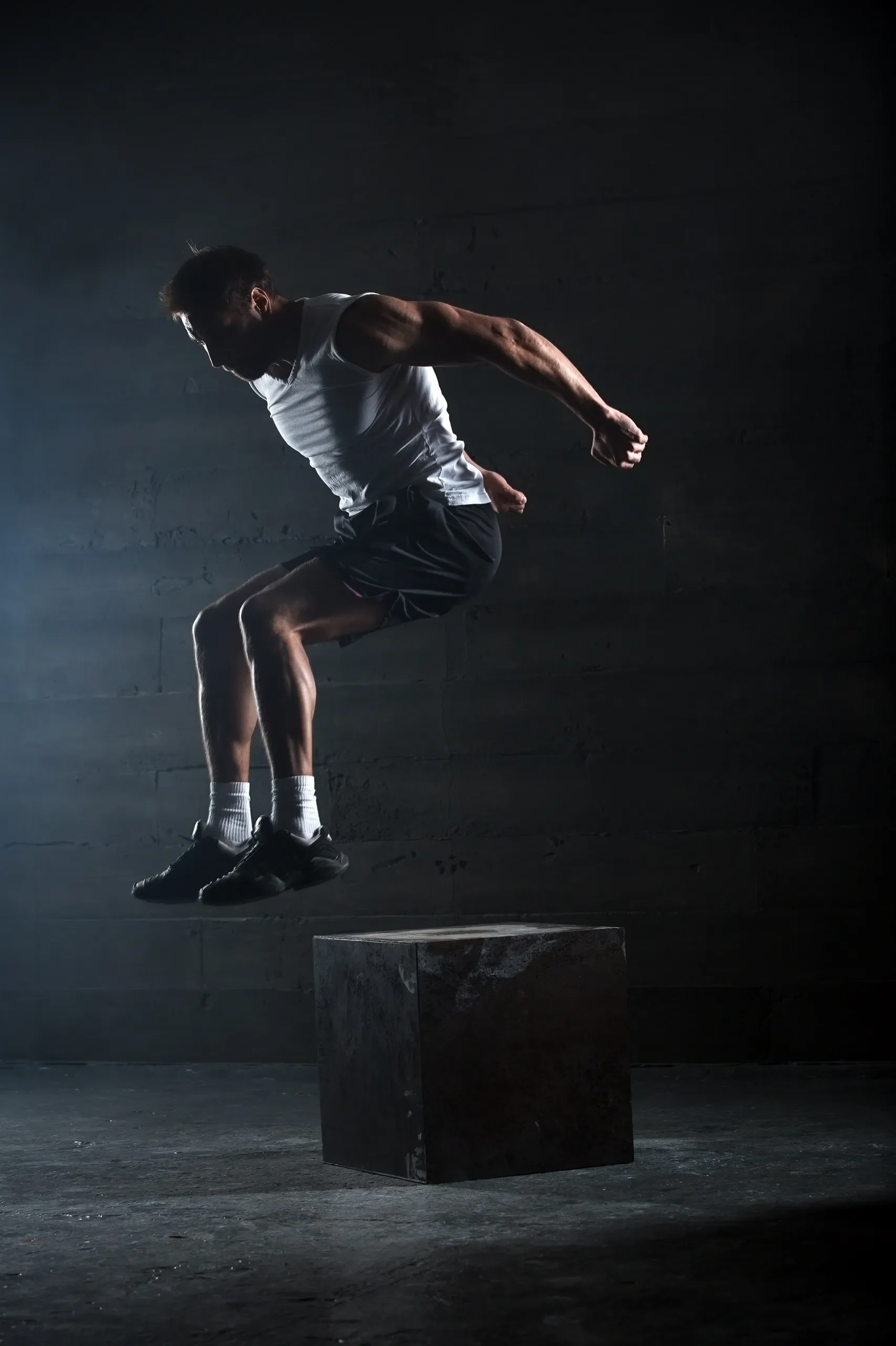 A man jumping from a black cube in a dark room, wearing a white sleeveless shirt, black shorts, white socks, and black shoes.