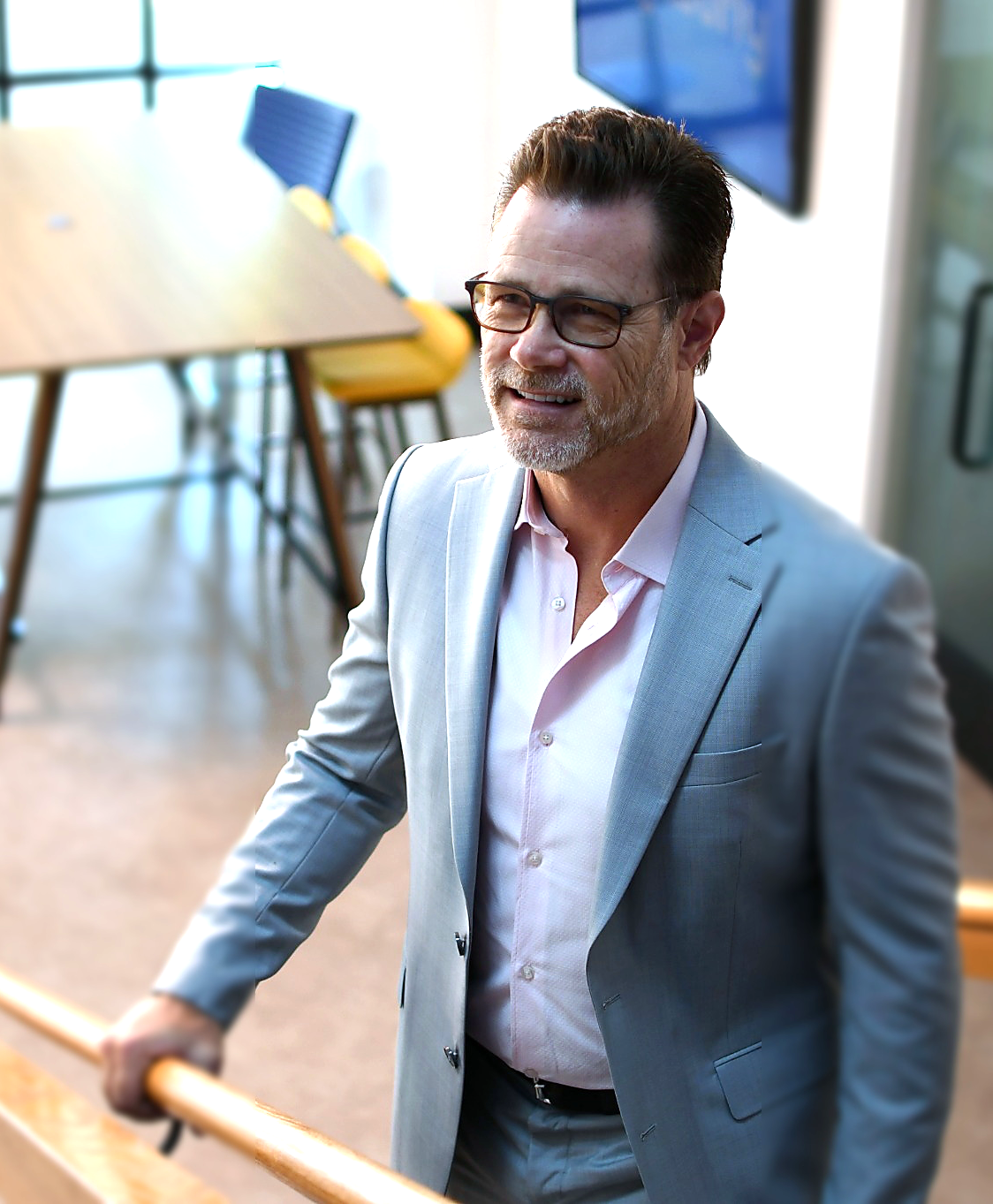 A middle-aged man with dark hair, glasses, and a beard, dressed in a light gray suit and pink shirt, standing and smiling in an office or conference room.