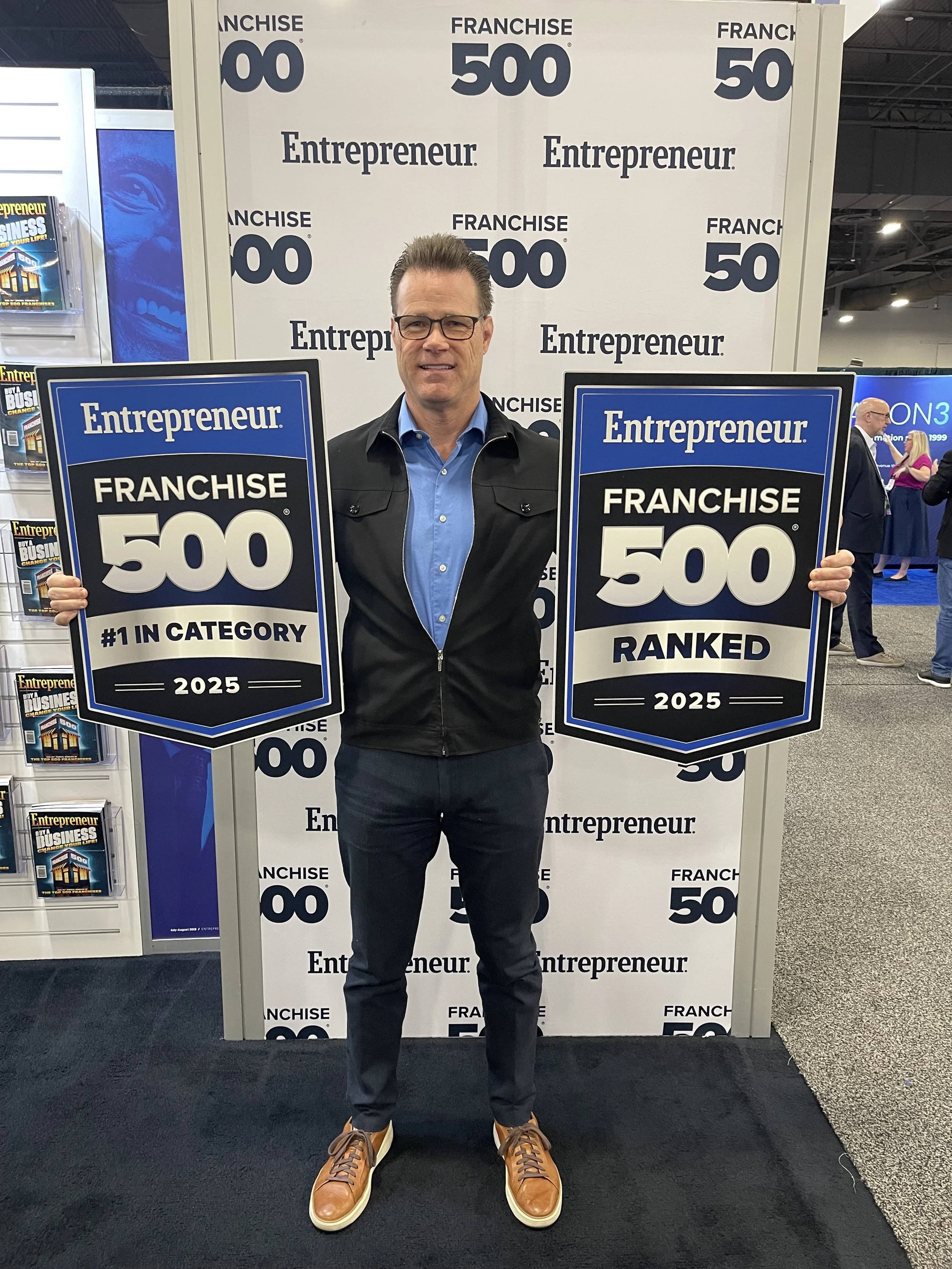 A man holding two large signs that read 'Entrepreneur Franchise 500', with one sign stating '#1 in Category 2025' and the other 'Ranked 2025', standing in front of a backdrop with repeated 'Franchise 500' and 'Entrepreneur' logos at a trade show or conference.