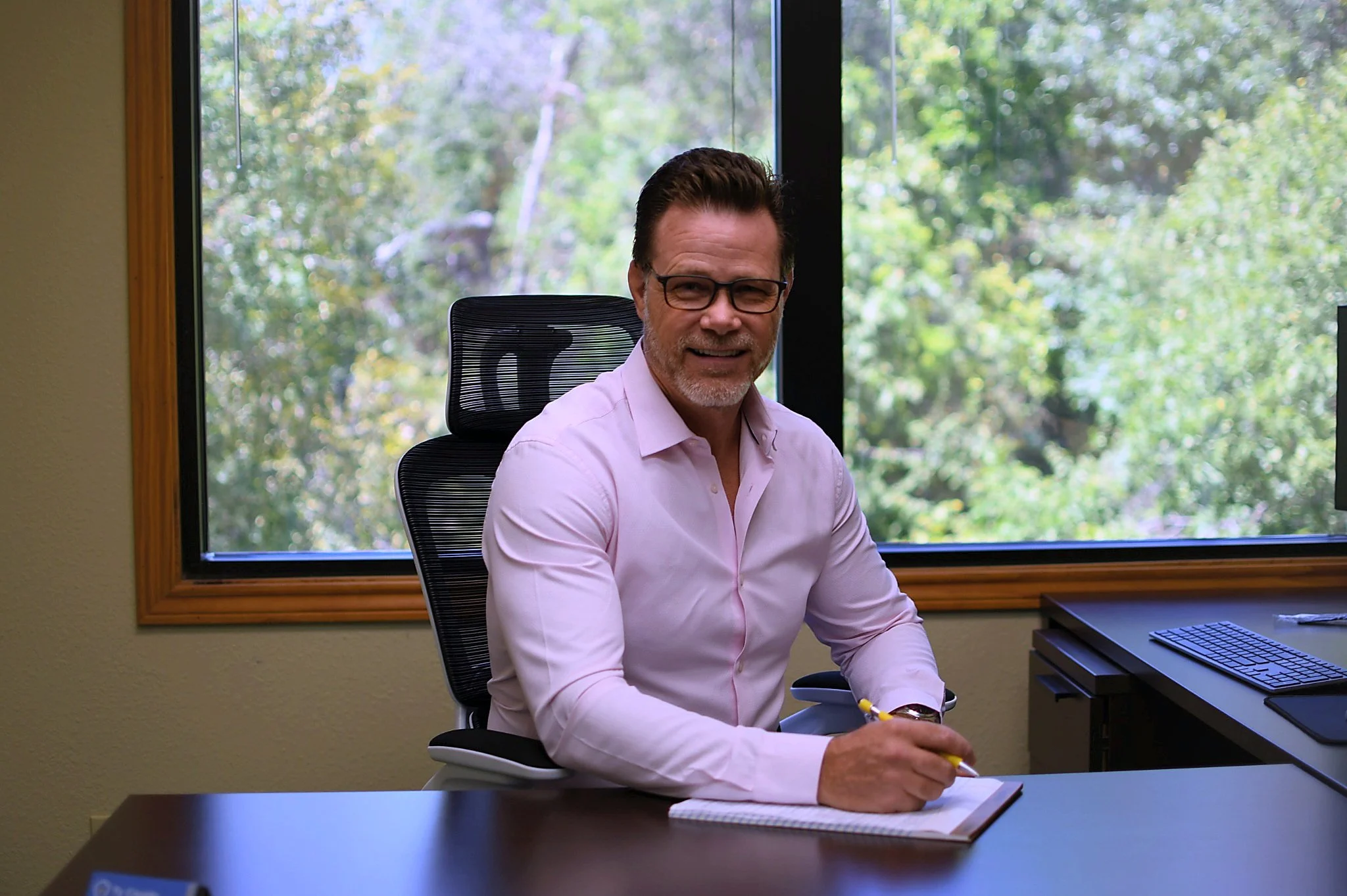 Man sitting at a desk in an office, smiling, wearing glasses and a pink shirt, with a notebook and pen, background of a large window with green trees outside.