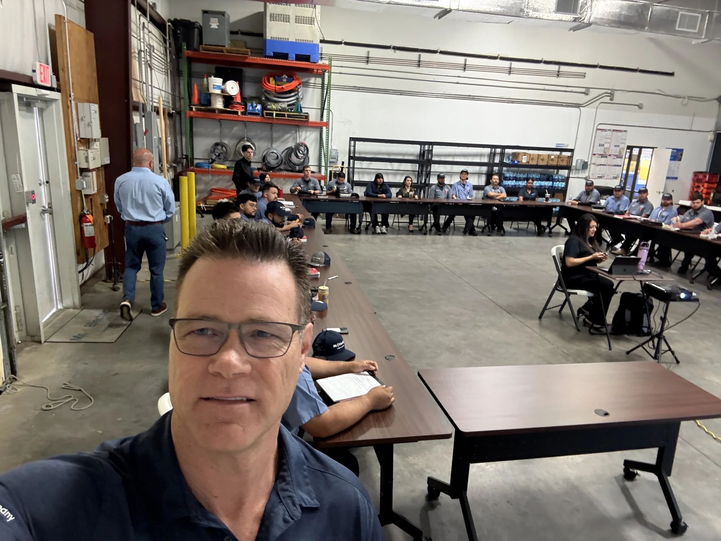 A man taking a selfie in a large industrial workspace with many people seated at tables forming a U-shape, some working on laptops, and large storage shelves filled with equipment in the background.