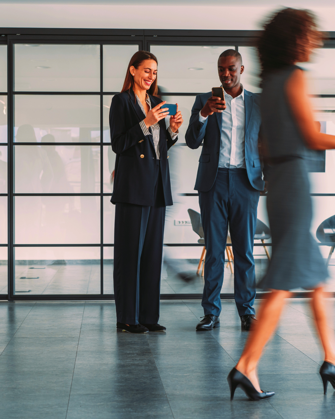 Two business professionals, a woman and a man, standing indoors using their smartphones, with another woman walking past them in the foreground.