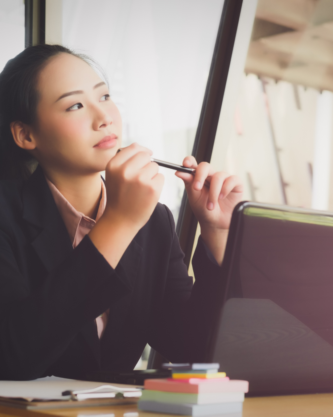 A young woman in a business suit sitting at a desk, holding a pen near her chin, looking thoughtful while working on a tablet or laptop, with a window in the background.