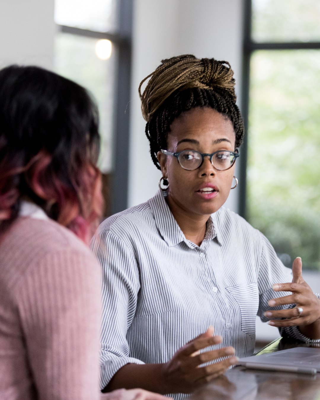 Two women engaged in conversation at a table in a well-lit room with large windows, one woman with glasses and braided hair speaking, the other woman facing away with pink and dark hair.