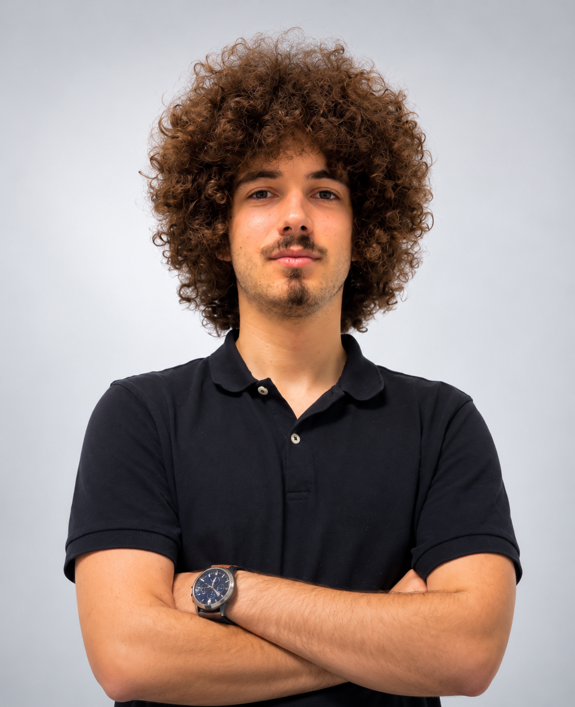 Homem jovem com cabelo cacheado volumoso, barba rala, camisa polo preta e braços cruzados, posando diante de fundo cinza claro.