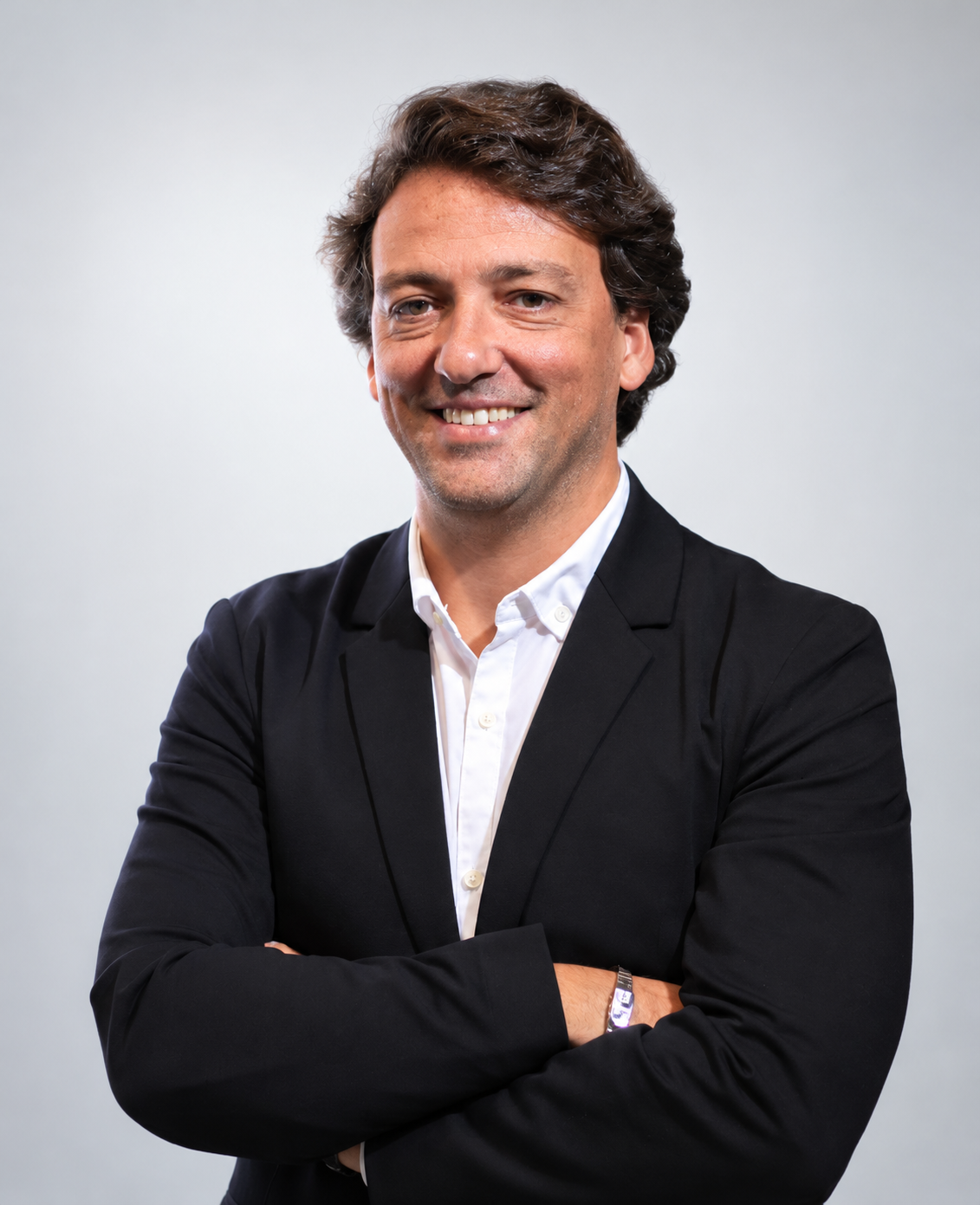 Retrato profissional de homem sorrindo, usando terno preto e camisa branca, com os braços cruzados sobre fundo cinza claro.