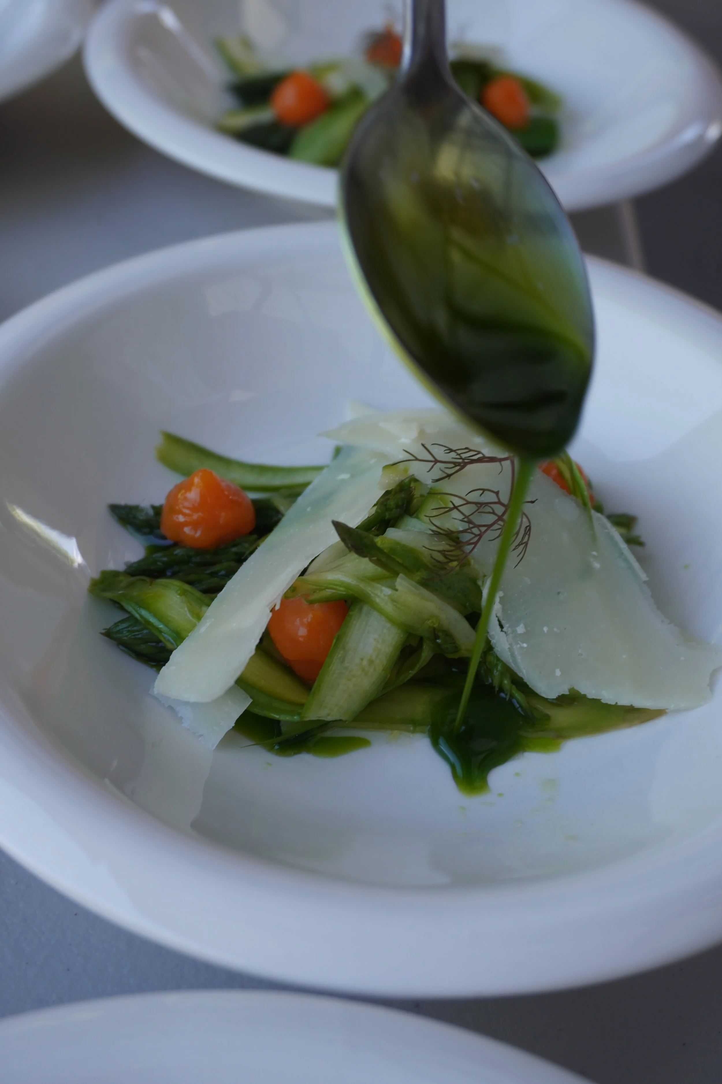 A white bowl containing a salad with julienned vegetables, cherry tomatoes, and shaved cheese, with a spoon resting inside.