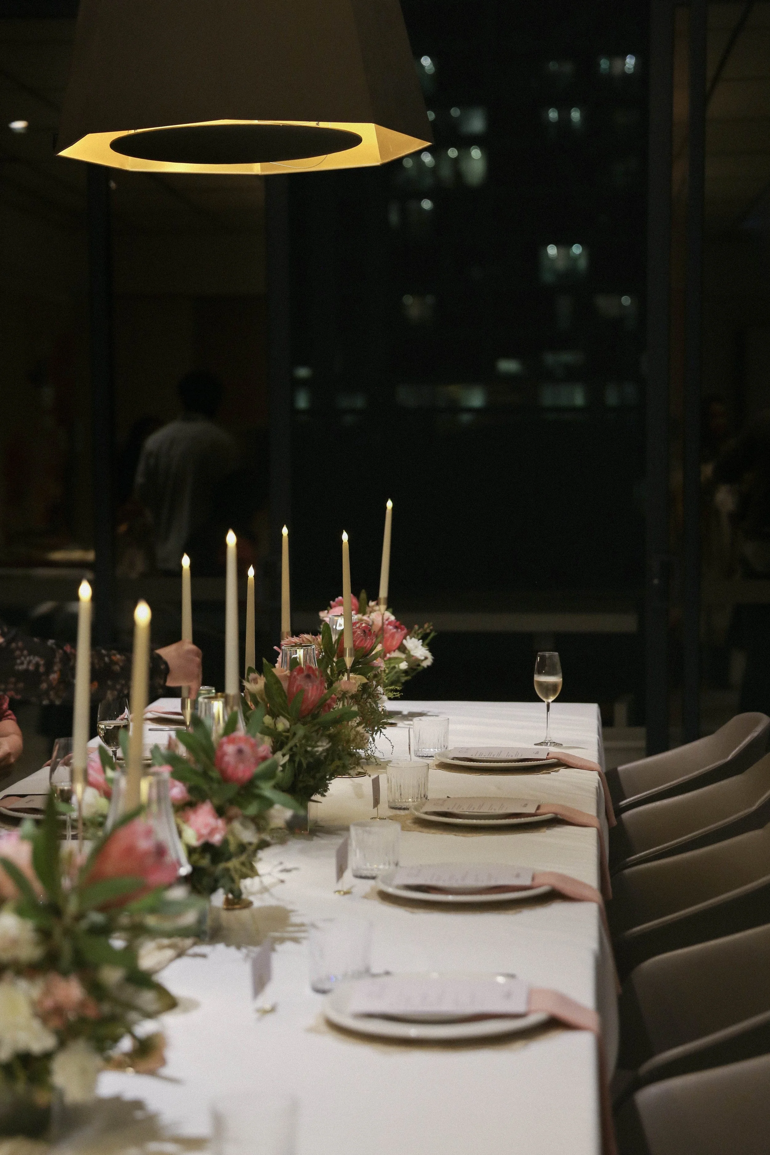 A long dining table set up for a private catered dinner party with floral center pieces, candles, and place settings, with a dark background and a hanging lamp above.
