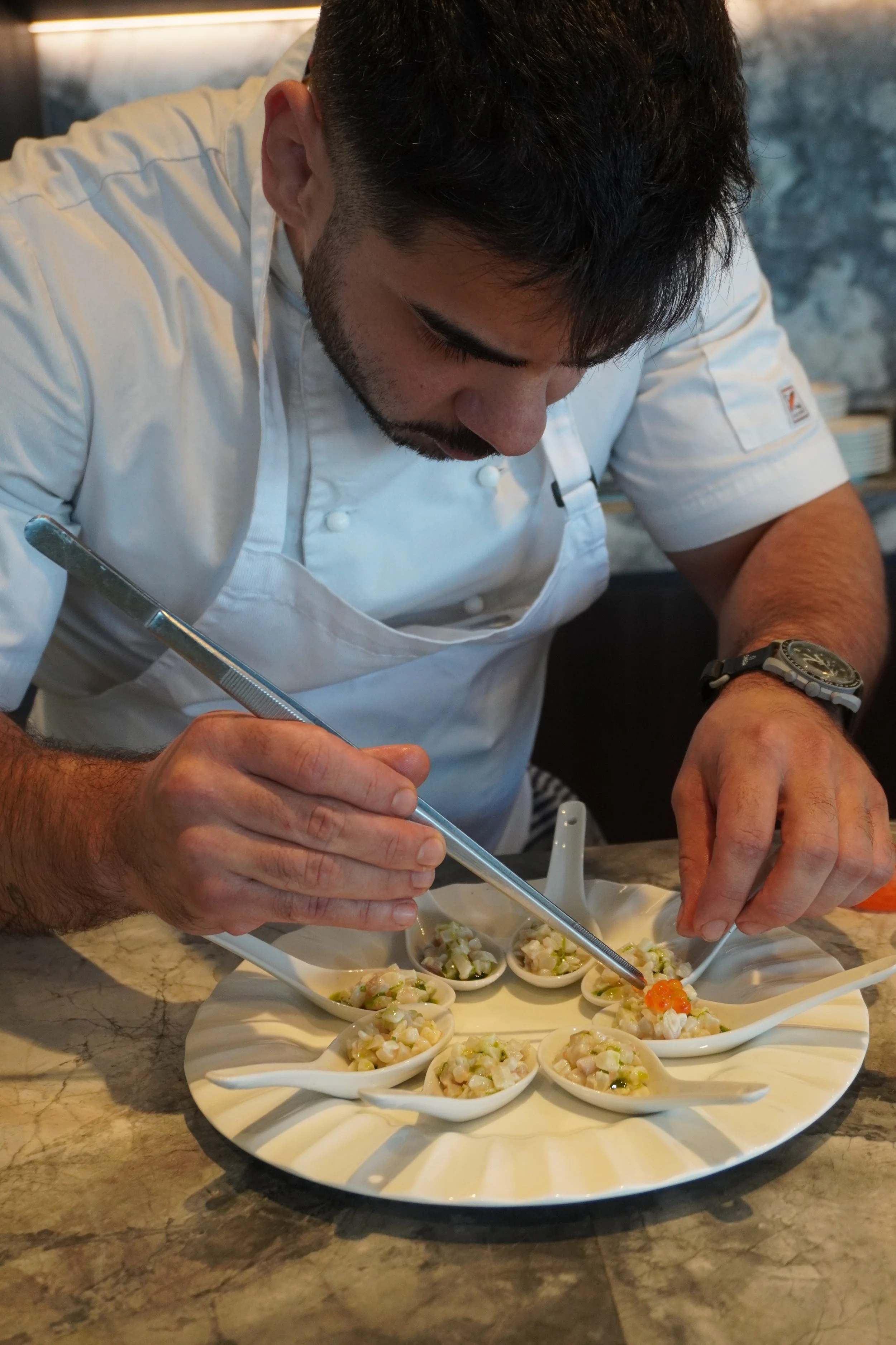 A chef in a white uniform preparing small dishes of food on a tray, using tweezers, in a kitchen setting.