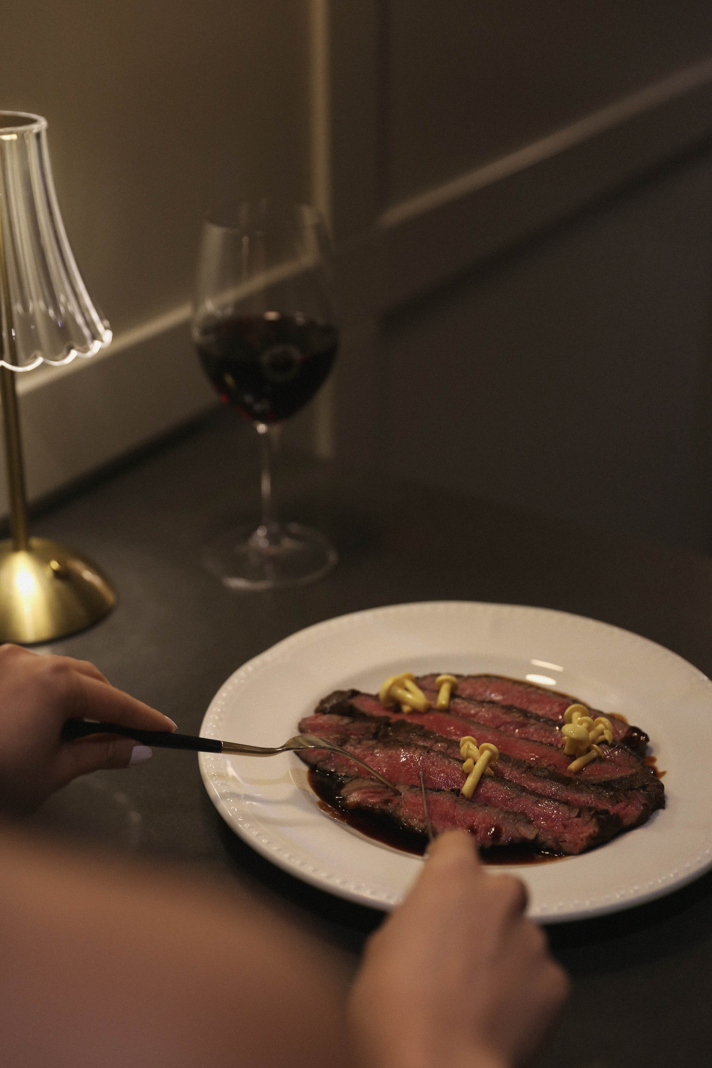 A dinner table with a plate of sliced steak, a glass of red wine, and a brass lamp in the background for a dinner party.