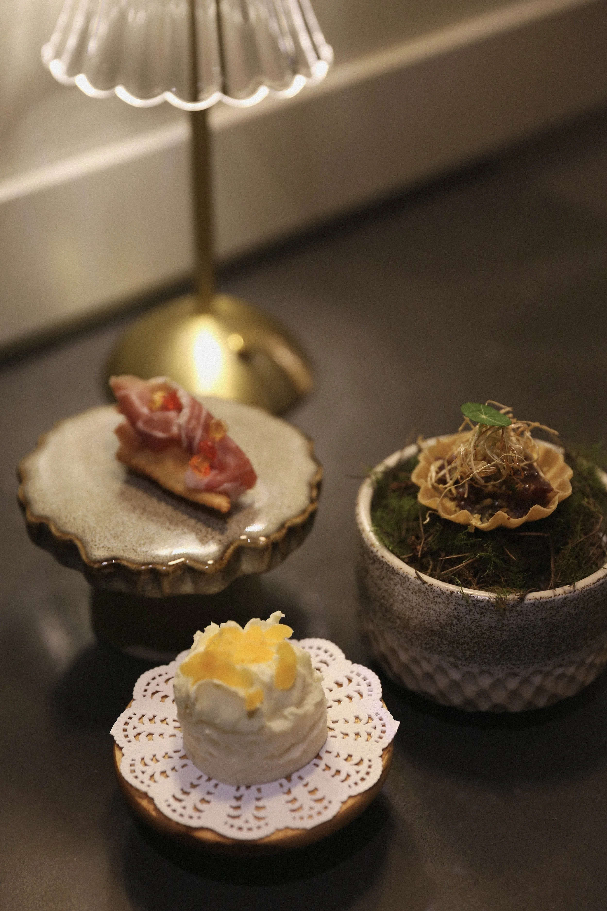 Assorted small canapés and snacks, including a puff pastry with toppings, a savoury tart with greens and sprouts, and a small pastry with a yellow topping, displayed on a black surface with a gold and silver lamp in the background.