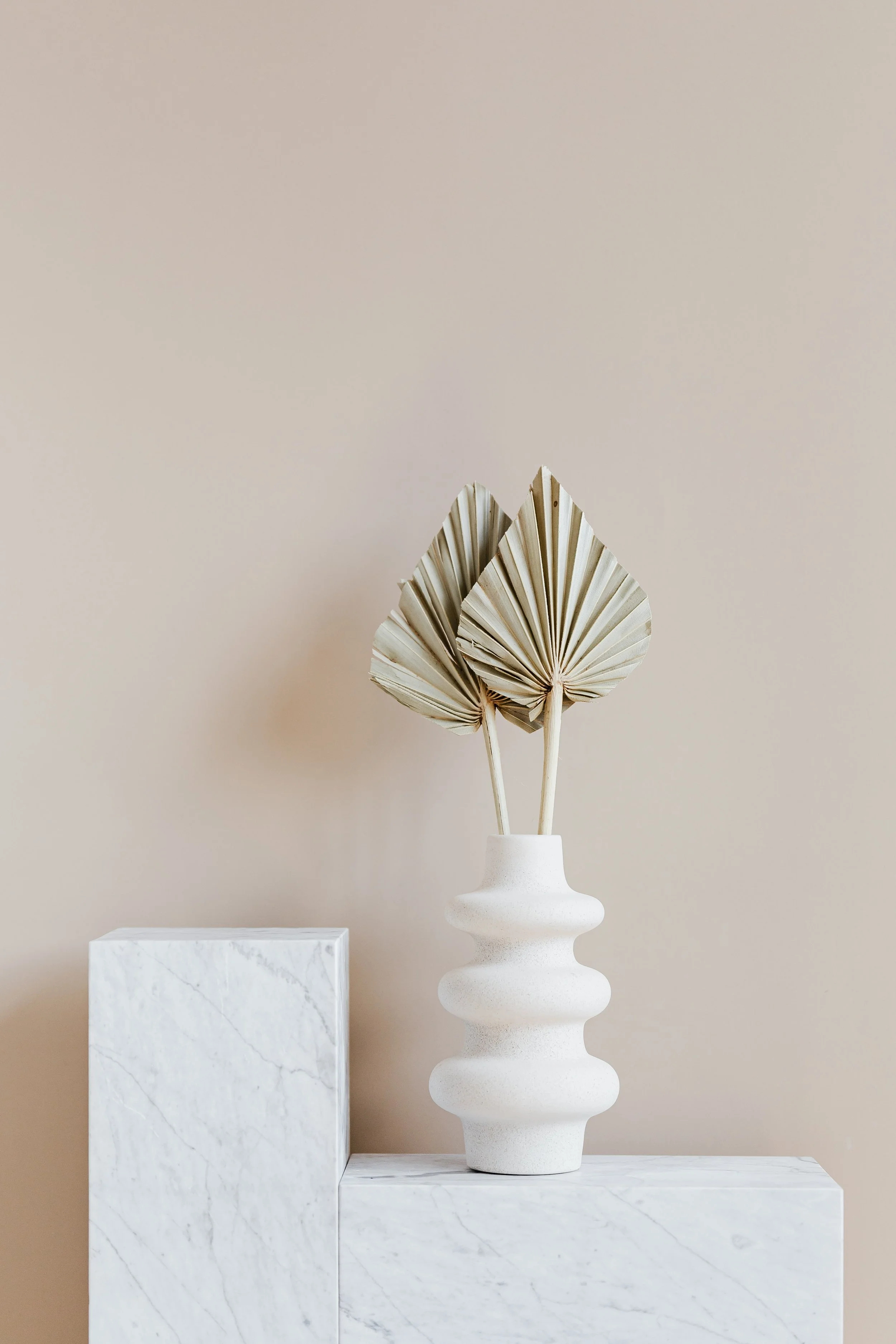 Decorative arrangement with dried fan palm leaves in a textured white vase on a white marble surface against a neutral beige wall.