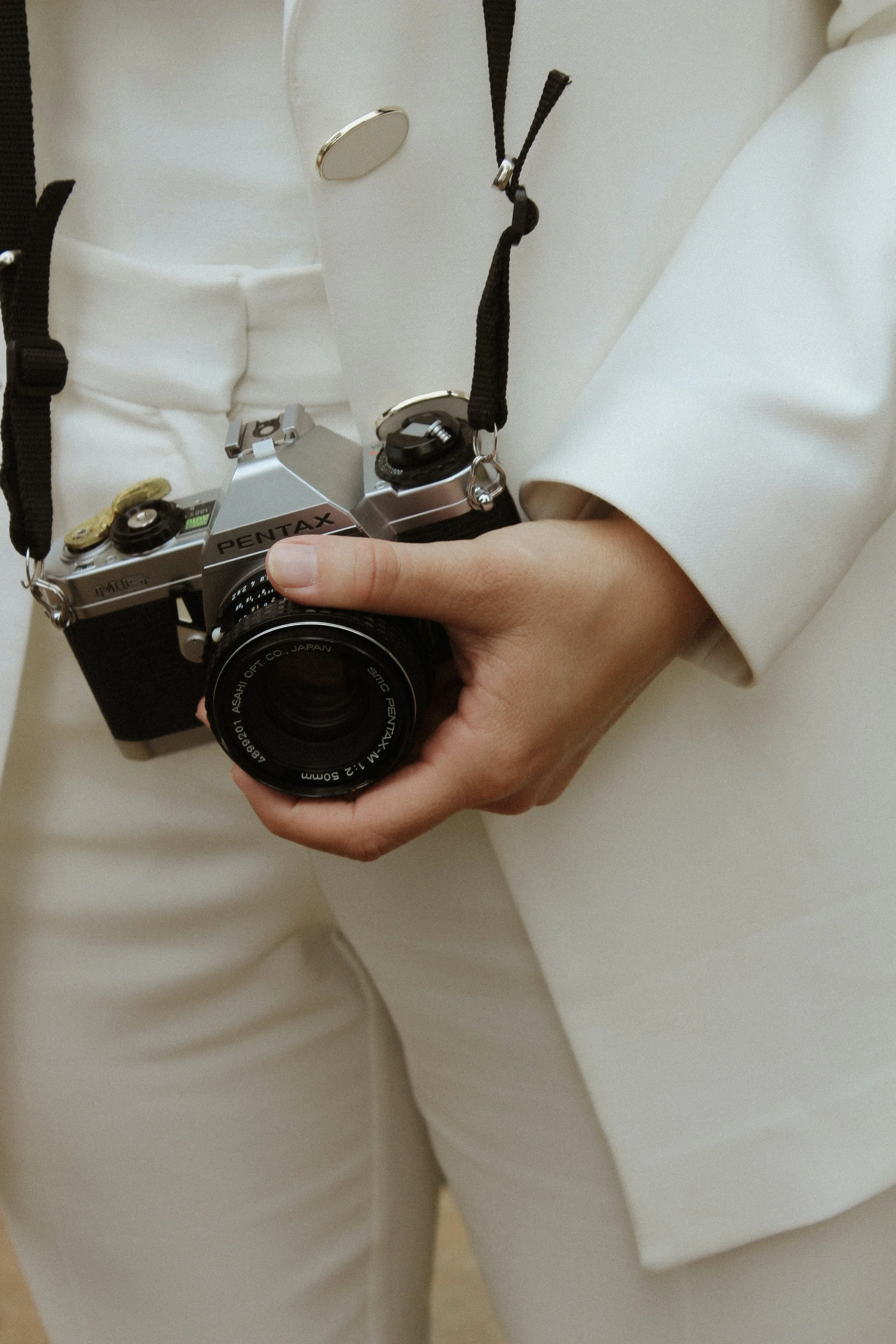 Person holding a silver Pentax film camera with black grip, dressed in beige suit.