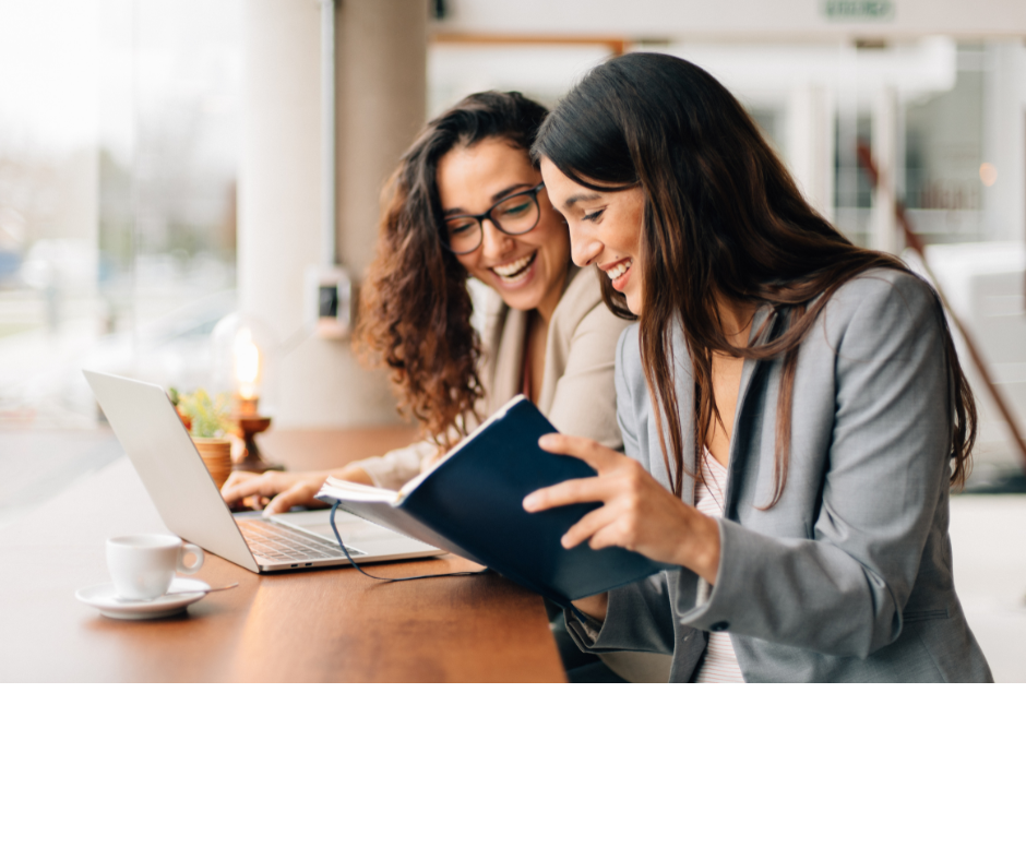 Two women sitting at a table in a cafe, one is looking at a notebook and the other is smiling and looking at the notebook, with a laptop and a coffee cup on the table.