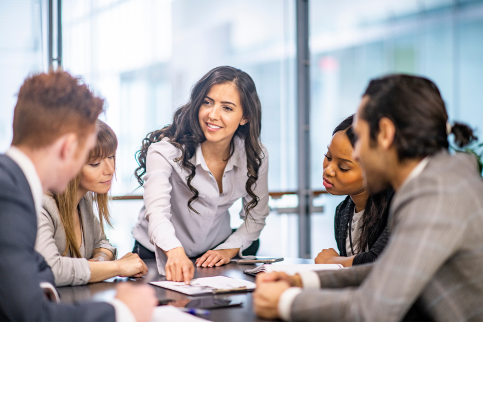 A diverse group of five professionals sitting around a conference table, engaged in a discussion, with a woman standing and speaking.